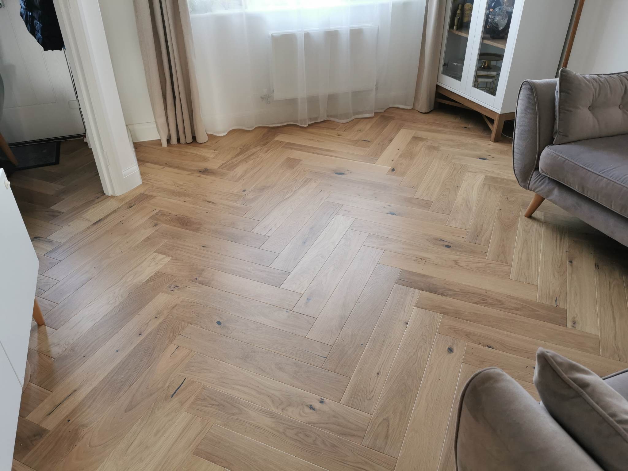 Parkiet ułożony w jodełkę francuską, z widocznymi naturalnymi przebarwieniami drewna, w jasnym wnętrzu z zasłonami i fragmentem sofy.