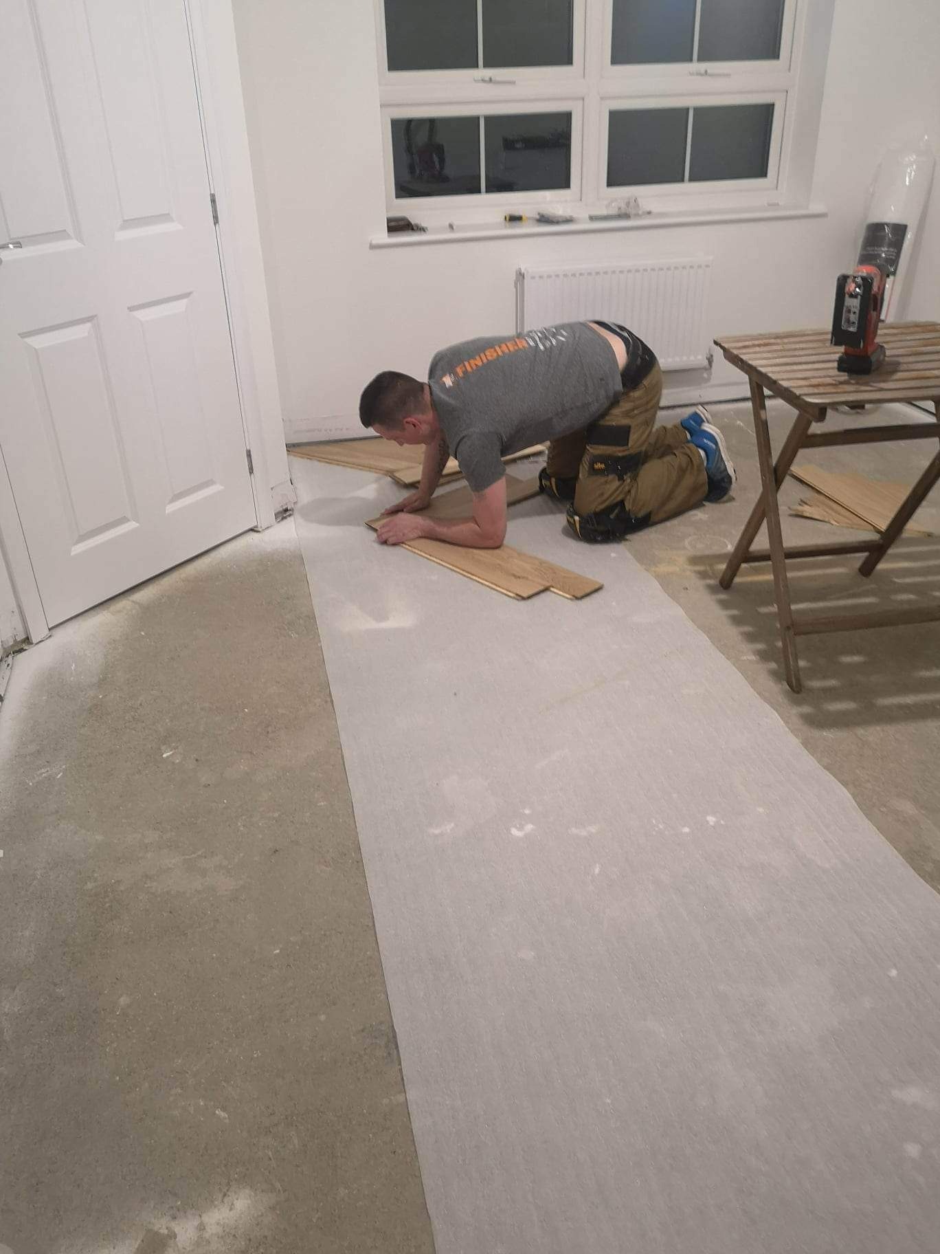 Parkieciarz na kolanach układa drewniane panele podłogowe na podkładzie w jasnym pokoju z oknem. Na stoliku narzędzia.