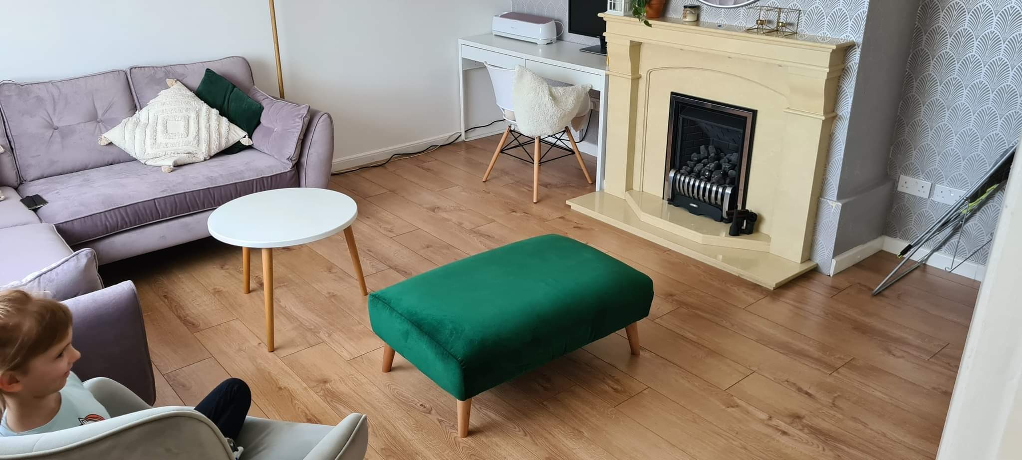 Salon z parkietem, kremowym kominkiem i zielonym pufem. Fioletowa sofa z poduszkami, biały stolik kawowy i biurko. Tapeta w stylu art deco. Widoczny fragment osoby.