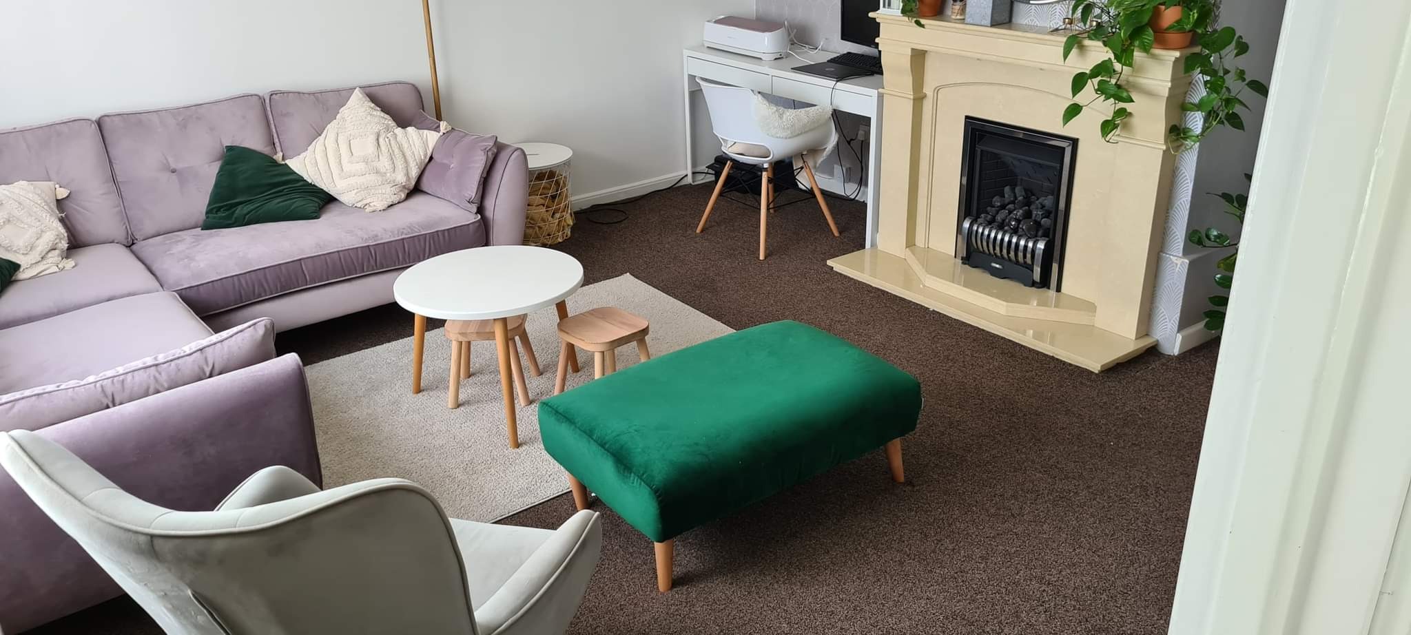 Jasny salon z liliową sofą, zielonym pufem i beżowym kominkiem. Na biurku laptop i drukarka. Wnętrze po remoncie, z elementami dekoracyjnymi i roślinami.