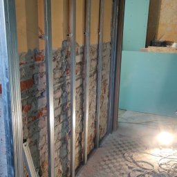 KP NOVA BUD Krzysztof Pisarek - Montaż metalowego stelażu na ceglanej ścianie, przygotowanie pod płyty gipsowo-kartonowe. Widoczny poziomica i przewody elektryczne na podłodze.