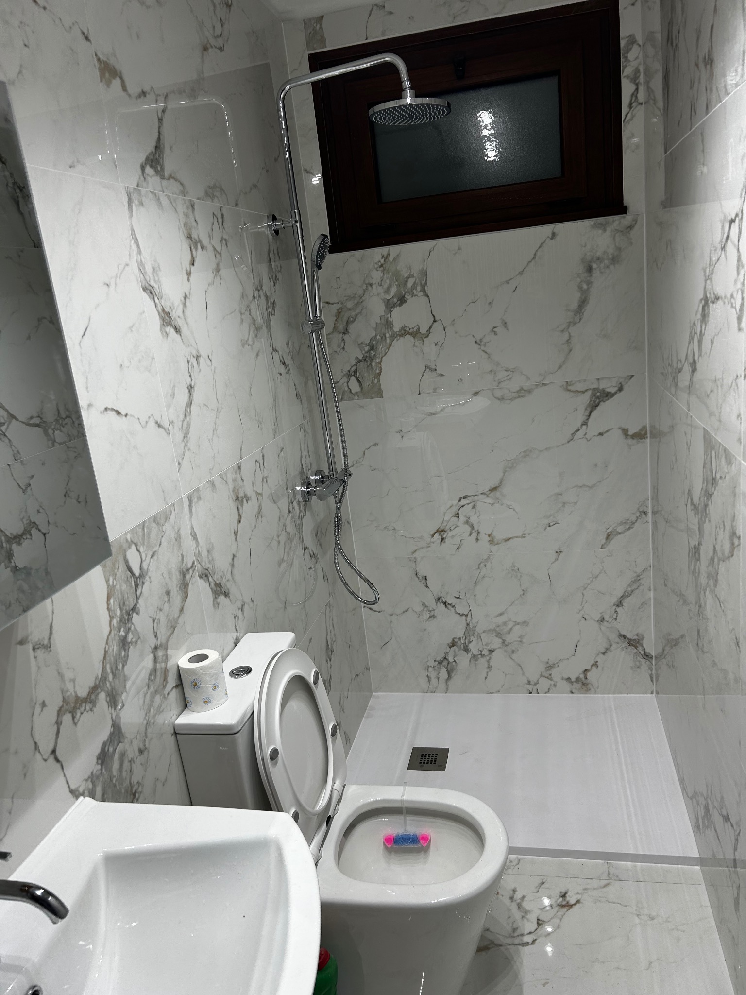 Wykończona łazienka z marmurowymi płytkami, kabiną prysznicową typu walk-in i nowoczesną armaturą. Uwagę zwraca okno w ciemnej ramie nad prysznicem.