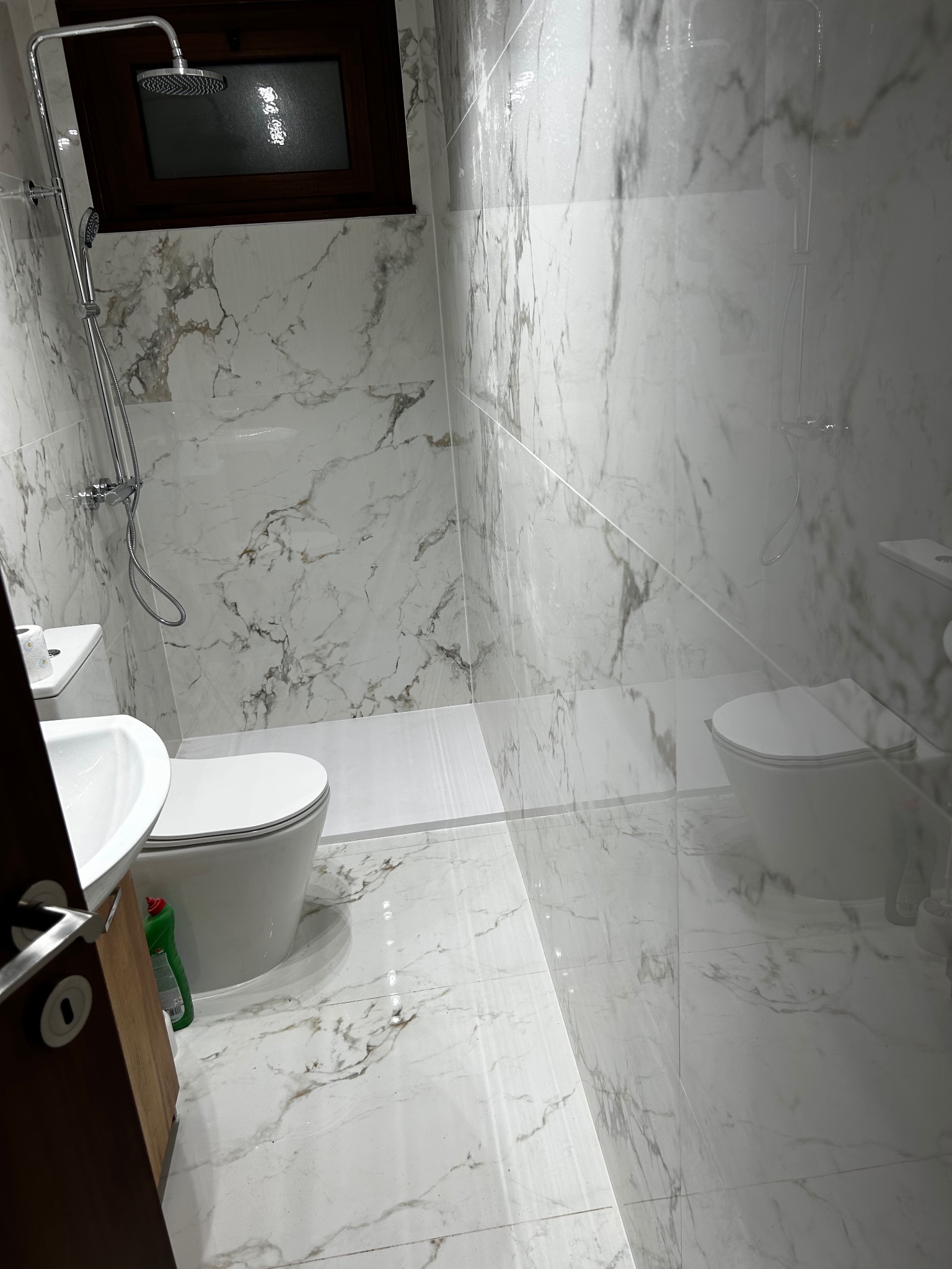 Biała łazienka z marmurowymi płytkami, kabiną prysznicową typu walk-in i wiszącą toaletą. Widoczny fragment umywalki i drzwi. Nowoczesny design z akcentem na czystość.