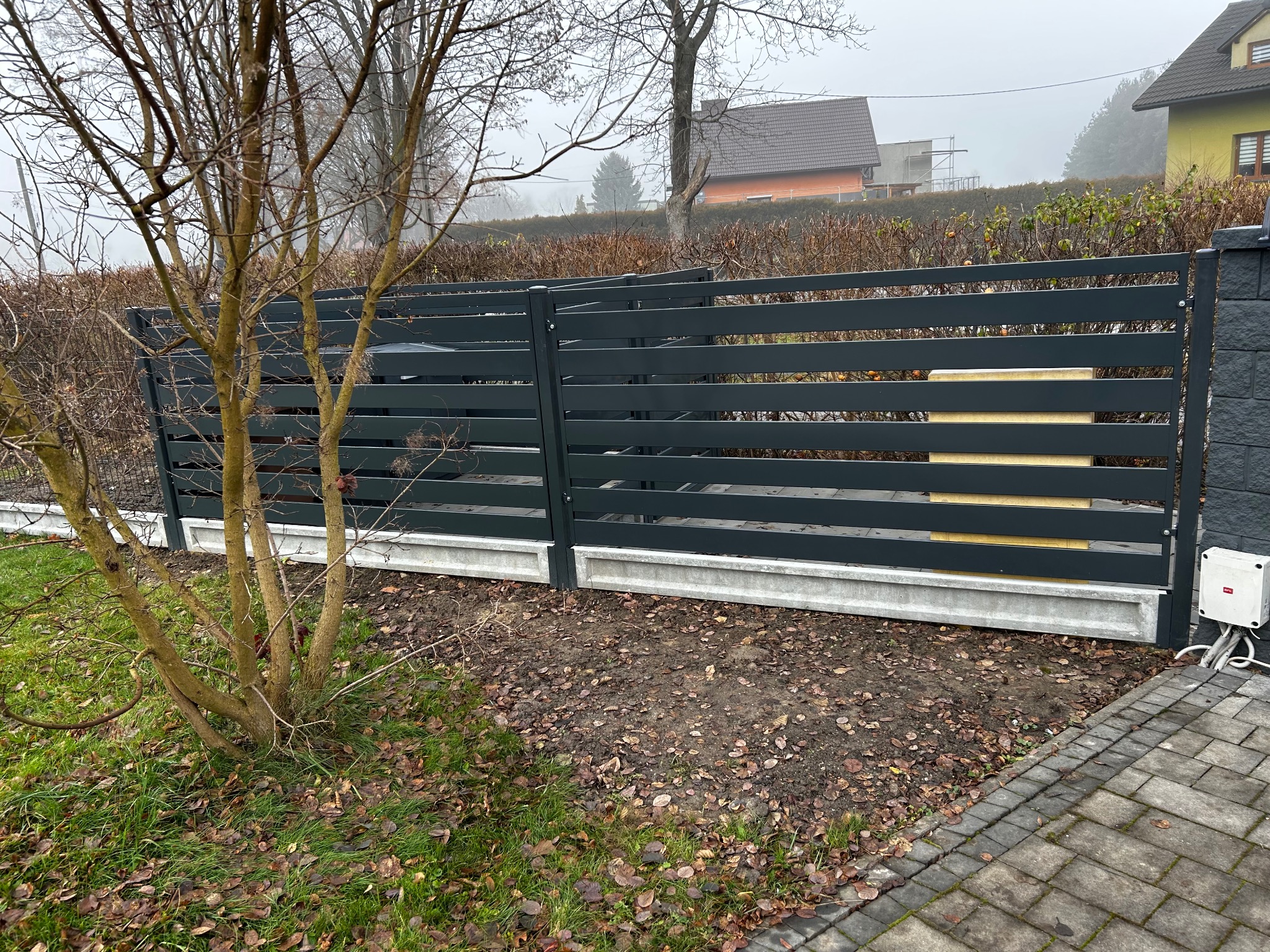 Nowoczesne ogrodzenie panelowe w kolorze grafitowym z podmurówką betonową, widoczne z perspektywy ogrodu, z drzewem na pierwszym planie i domami w tle.
