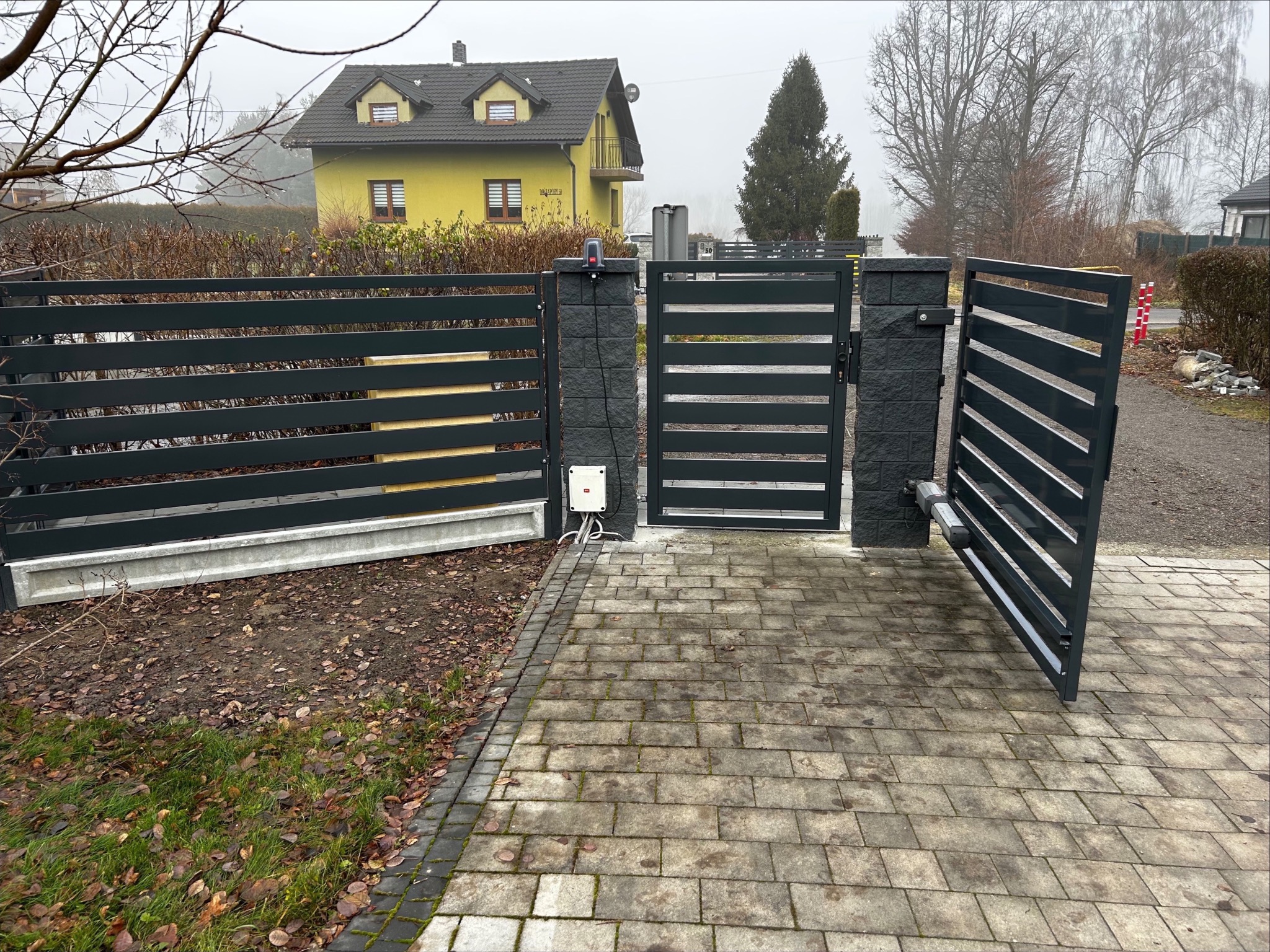 Nowoczesne ogrodzenie panelowe z automatyczną bramą wjazdową, widoczne z zewnątrz posesji. Szara kolorystyka, poziome panele, kostka brukowa na podjeździe, dom w tle.
