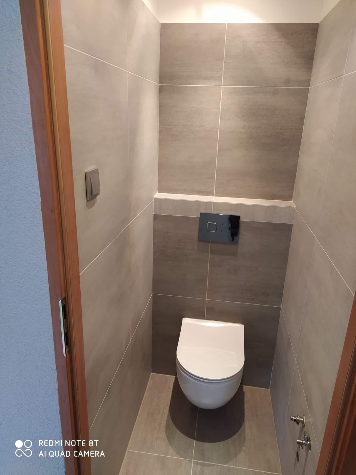 Nowoczesna toaleta w szarościach: płytki imitujące beton, podwieszana miska WC, chromowany przycisk spłukujący. Minimalistyczny design, subtelne oświetlenie.