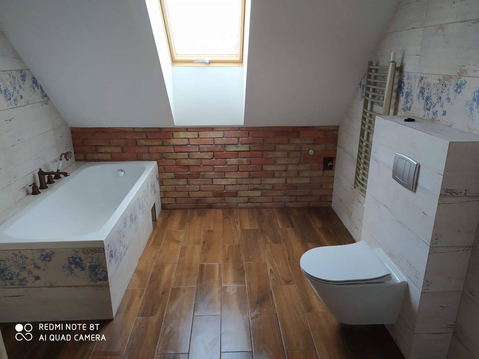 Wykończona łazienka na poddaszu z oknem dachowym. Brązowe płytki podłogowe imitujące drewno, biała wanna z dekorami, toaleta podwieszana i ściana z cegły.