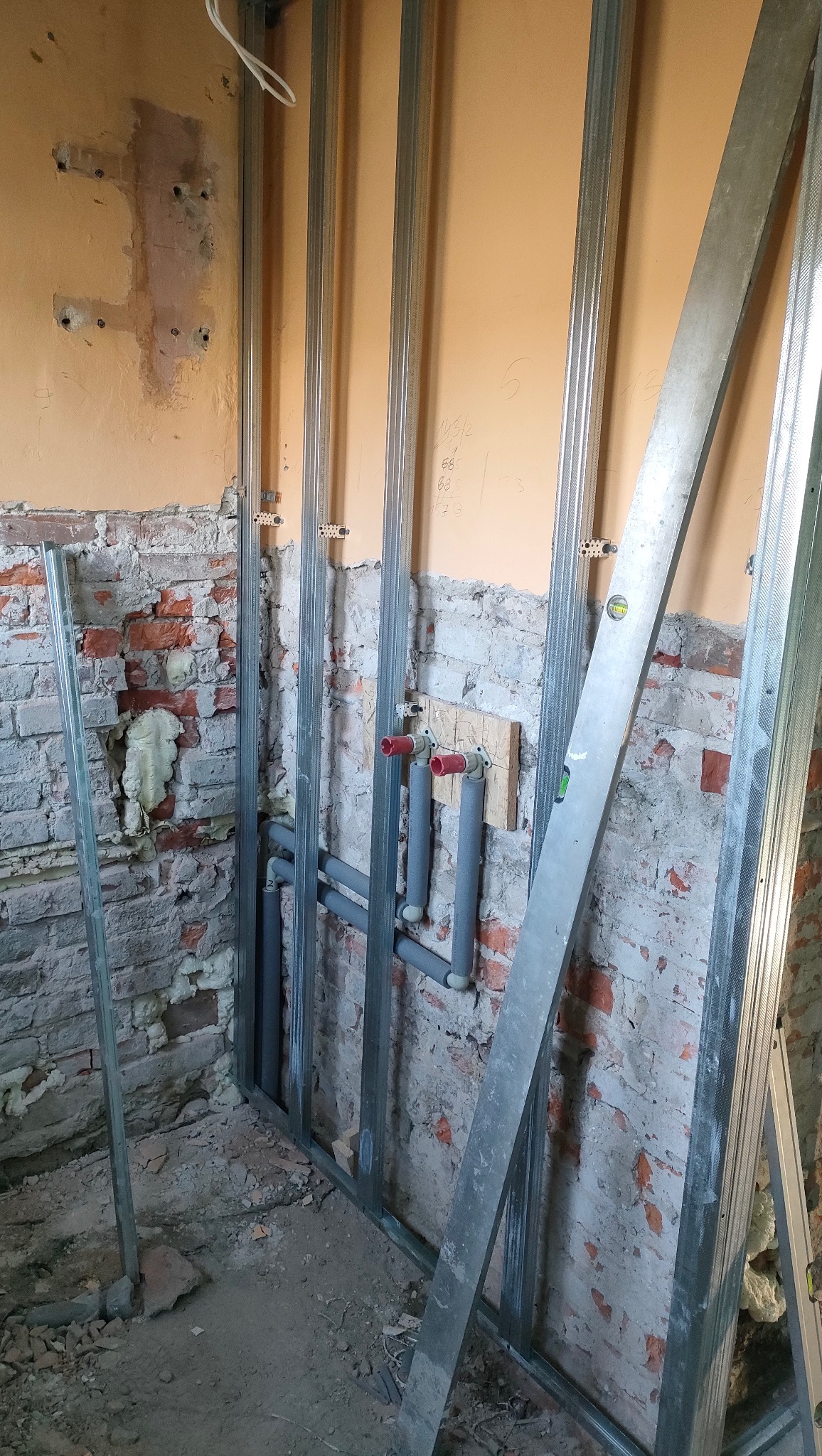 Stelaż z profili stalowych przy starej, częściowo obitej ścianie z cegły i instalacją wodną. Widoczny poziomica. Prace przygotowawcze do wykończenia wnętrza.