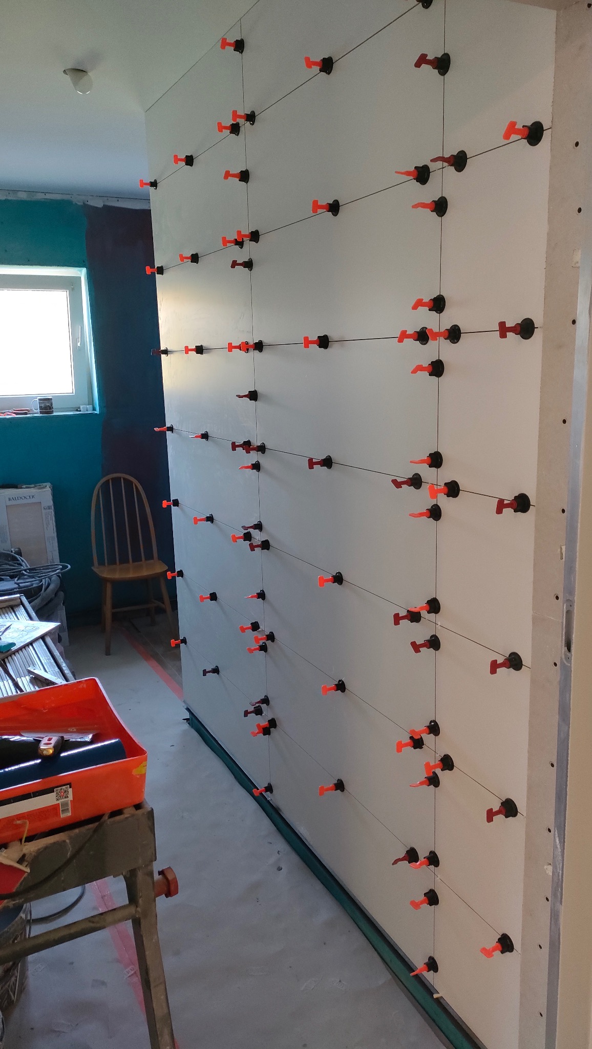 Ściana z płyt kartonowo-gipsowych, na której zamontowano system poziomowania płytek z pomarańczowymi klipsami, przygotowana do dalszych prac wykończeniowych.
