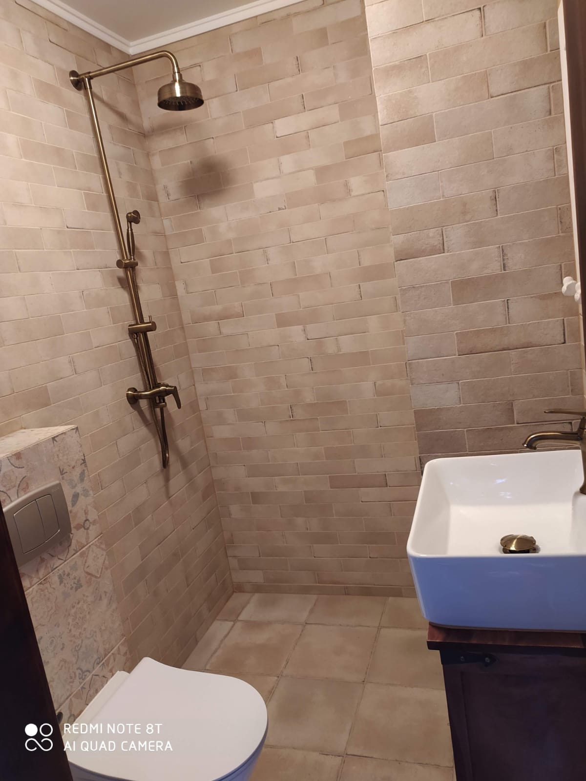 Beżowa łazienka z armaturą w stylu retro: bateria umywalkowa i deszczownica w kolorze starego złota, płytki imitujące cegłę, biała umywalka nablatowa i toaleta.
