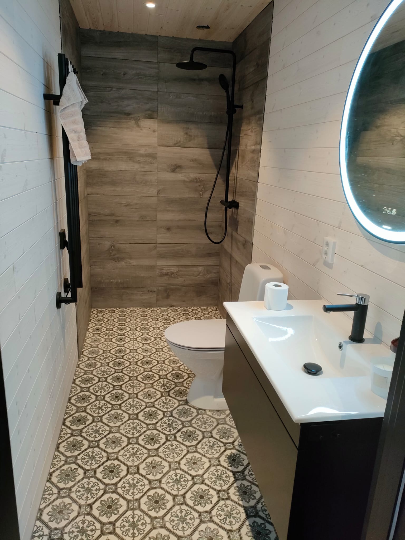 Wykończona łazienka w Zabrzegu: białe panele ścienne, płytki imitujące drewno pod prysznicem, wzorzysta podłoga, czarna armatura i okrągłe lustro z podświetleniem.