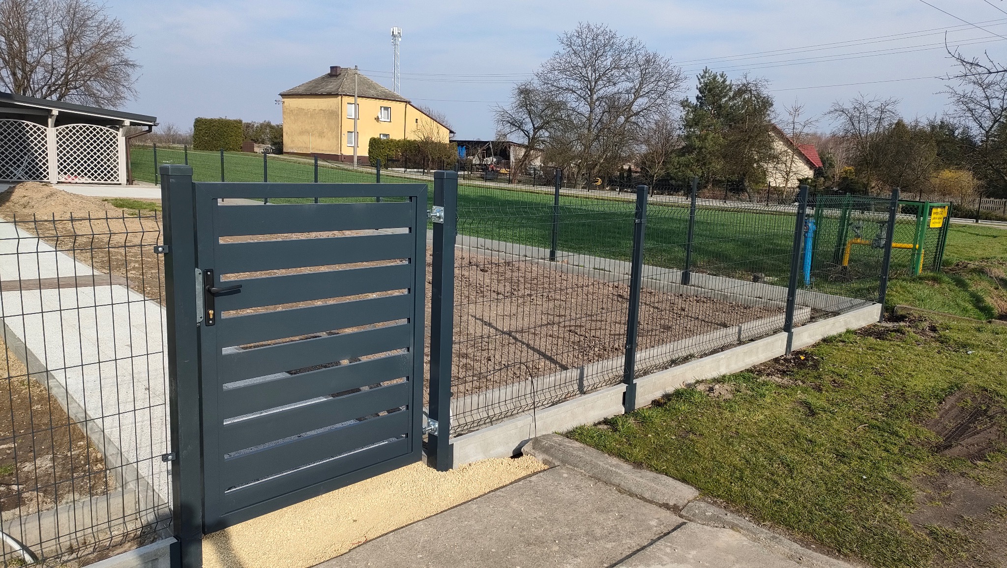 Nowoczesna, grafitowa brama lamelowa z furtką, część ogrodzenia panelowego z podmurówką, na tle wiejskiego krajobrazu z domami i drzewami.