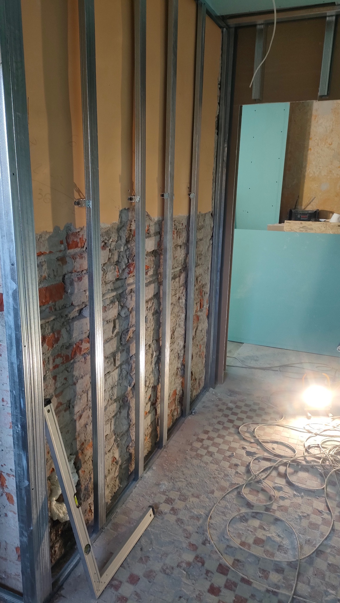 Montaż metalowego stelażu na ceglanej ścianie, przygotowanie pod płyty gipsowo-kartonowe. Widoczny poziomica i przewody elektryczne na podłodze.