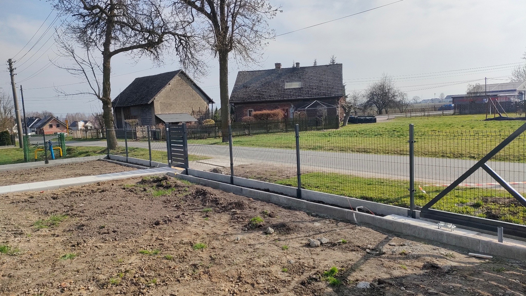 Nowoczesne ogrodzenie panelowe z bramką, osadzone na betonowej podmurówce, oddziela posesję od drogi. Widoczne budynki w tle i przygotowany teren pod ogród.