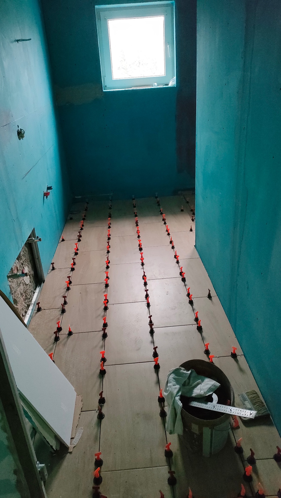 Układanie płytek podłogowych w łazience z użyciem systemu poziomowania; widoczne narzędzia i wiadro. Ściany przygotowane pod dalsze prace wykończeniowe.