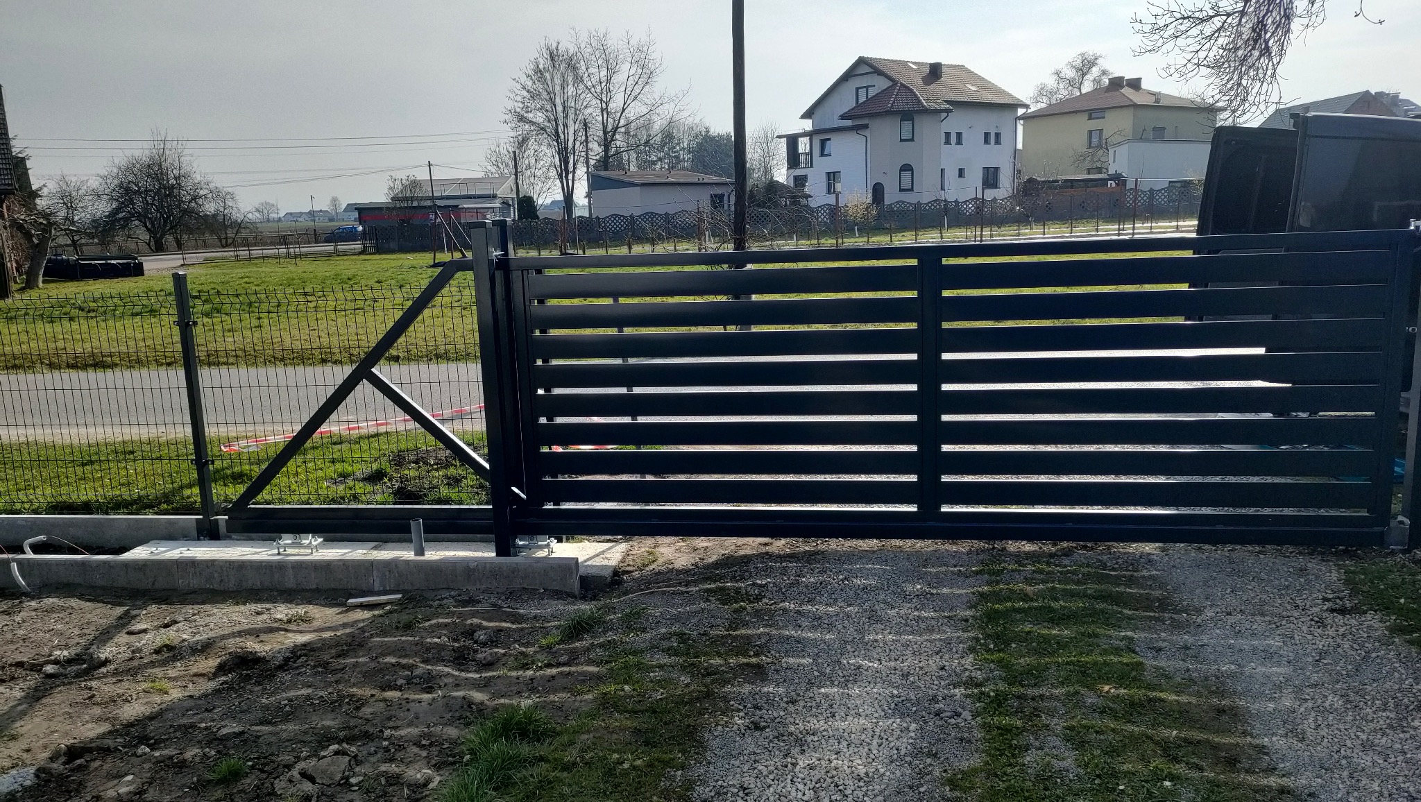 Nowoczesna, pozioma brama przesuwna w kolorze antracytowym, zamontowana na betonowej podstawie. W tle widoczny dom jednorodzinny i ogrodzenie z siatki.