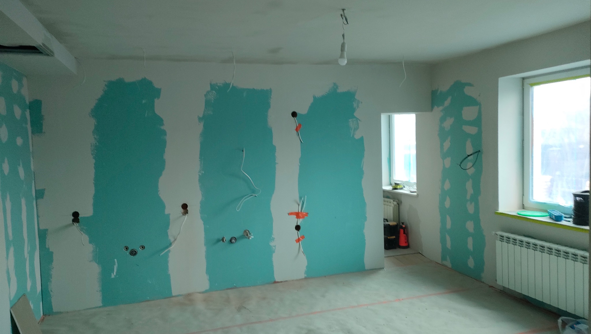 Ściany w trakcie szpachlowania i przygotowywania pod malowanie, widoczne instalacje elektryczne i hydrauliczne. Fragment grzejnika i okna w tle.