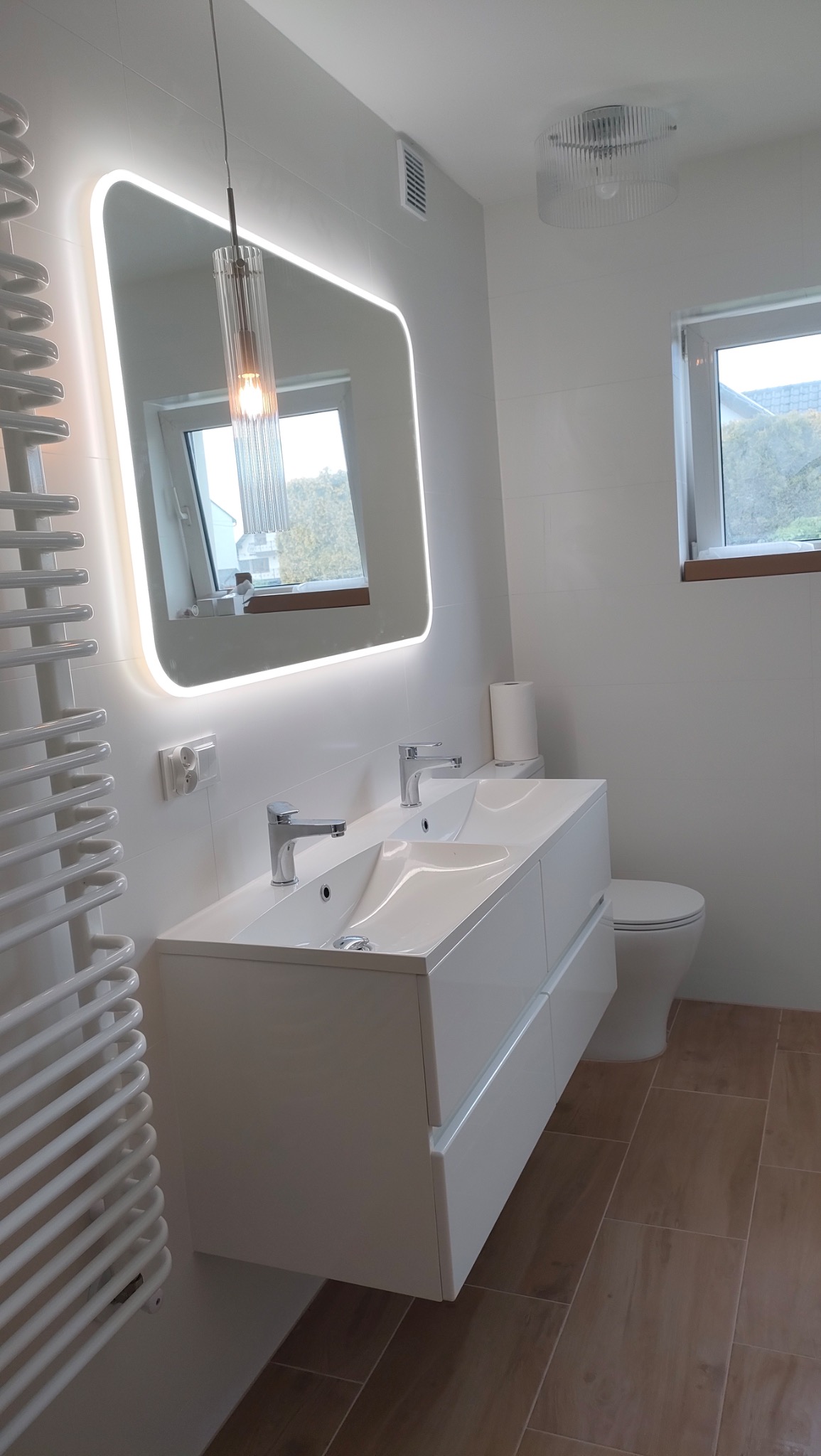 Nowoczesna łazienka z podwójną umywalką, lustrem z podświetleniem LED i grzejnikiem drabinkowym. Brązowe płytki podłogowe i białe ściany tworzą elegancki kontrast.