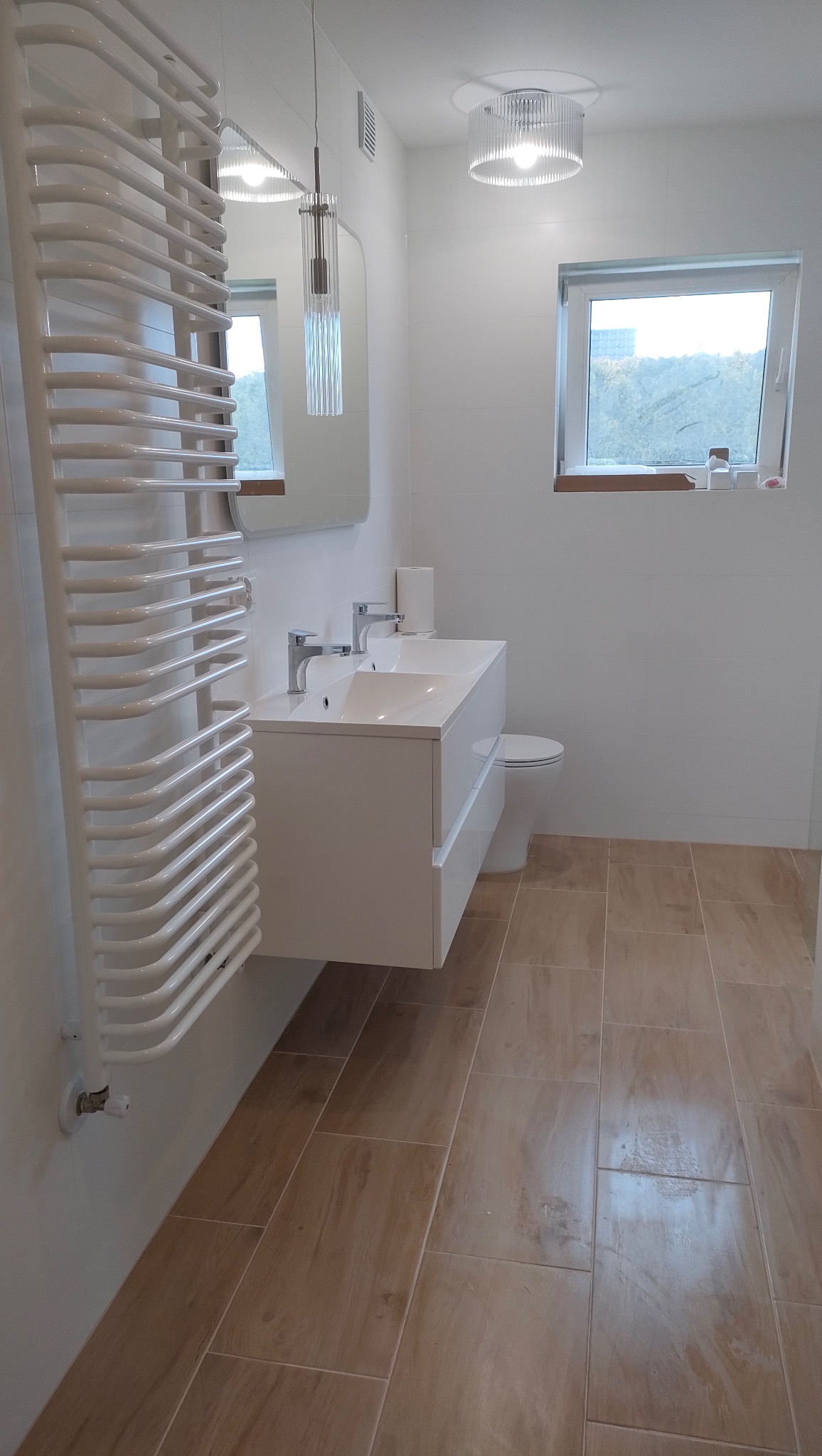 Jasna łazienka po remoncie: beżowe płytki podłogowe, biała umywalka z szafką, grzejnik drabinkowy, lustro i okno. Minimalistyczny design, czyste linie.