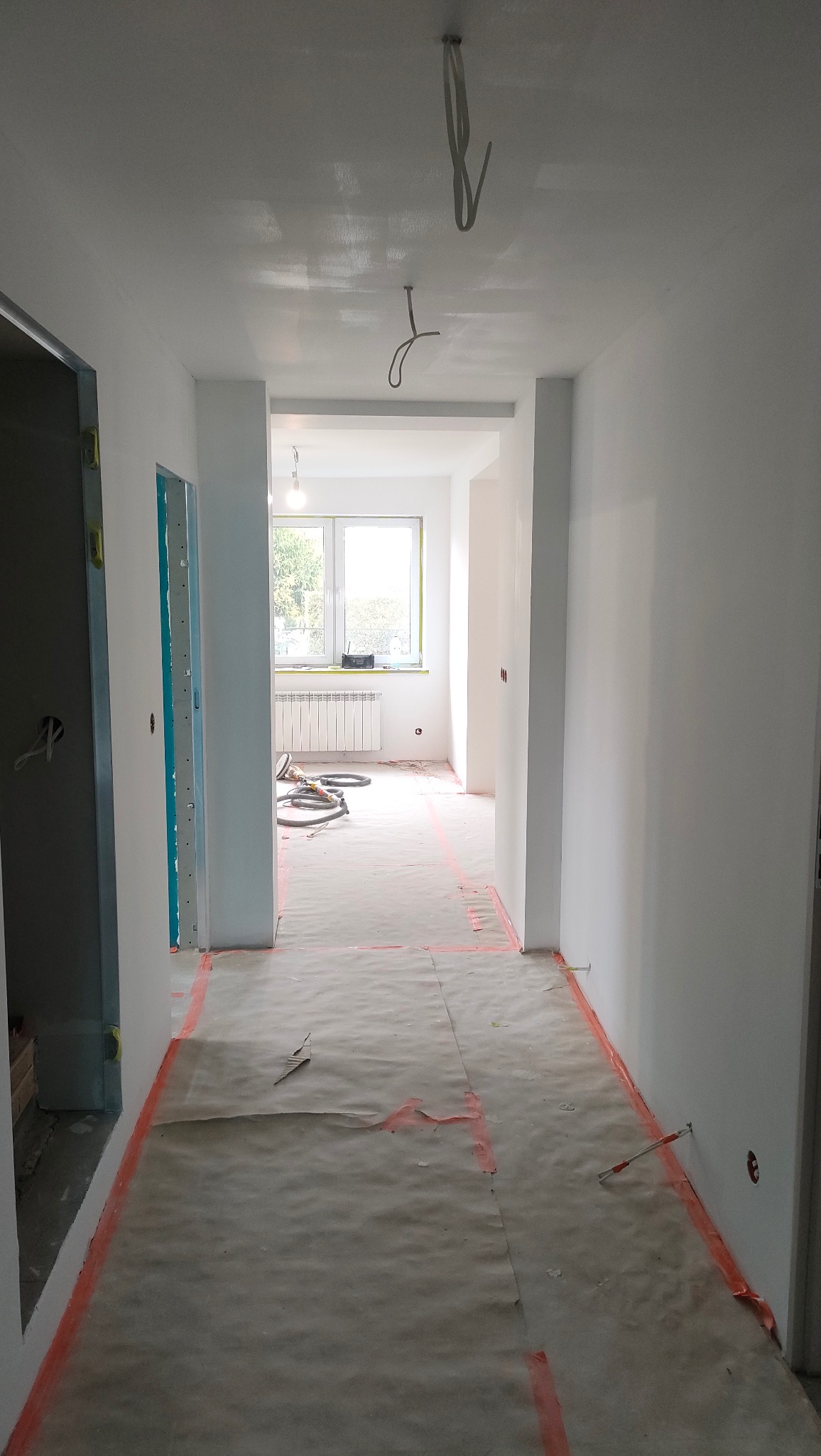 Białe, odświeżone wnętrze mieszkania w trakcie malowania. Podłoga zabezpieczona papierem i taśmą. Widoczne kable i grzejnik. Jasne, naturalne światło.
