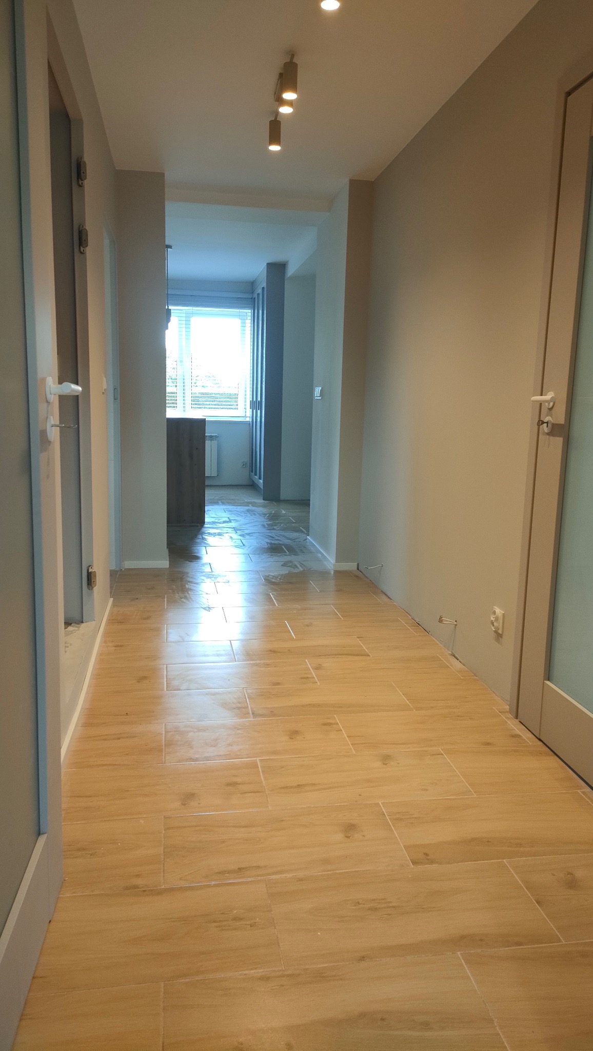 Jasne, drewnopodobne płytki podłogowe w korytarzu, widok w głąb mieszkania. Ściany w odcieniach beżu, sufit z oświetleniem punktowym. Częściowo widoczne drzwi po bokach.