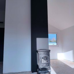 Maciej Arent - Poddasza i wykończenia - Wnętrze po remoncie: białe ściany, czarny filar, ozdobny piec żeliwny. Podłoga przygotowana do dalszych prac wykończeniowych. Okno z widokiem na zewnątrz.