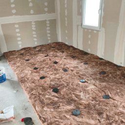 Maciej Arent - Poddasza i wykończenia - Wnętrze w trakcie prac: wełna mineralna z czarnymi mocowaniami na podłodze, ściany z płyt kartonowo-gipsowych przed wykończeniem, okno. Widoczny pojemnik z klejem.