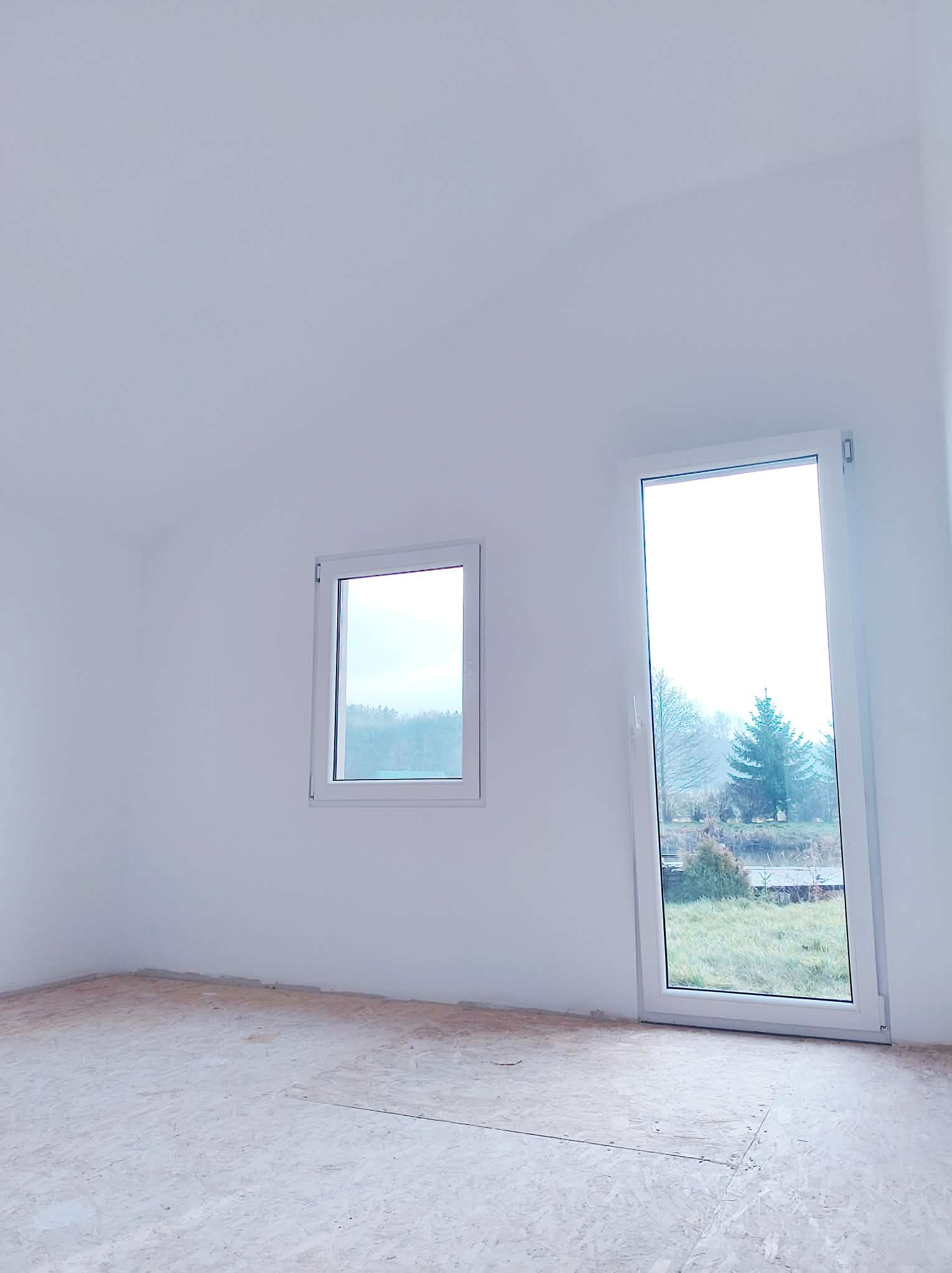 Jasne wnętrze z dwoma oknami o różnych rozmiarach, surowe ściany i podłoga z płyt OSB. Widok na zewnątrz przez okna.