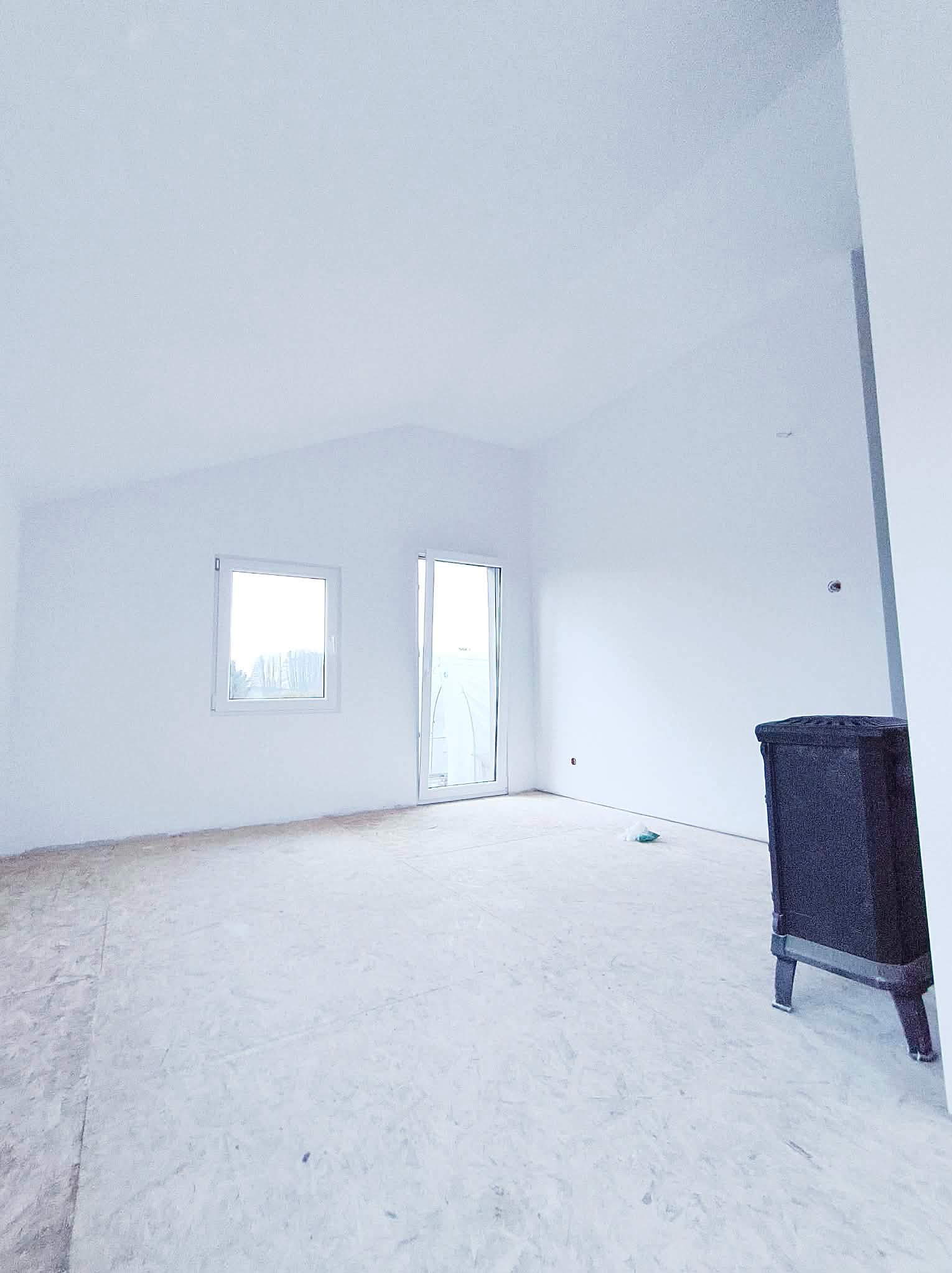 Jasne wnętrze z oknem i drzwiami balkonowymi po malowaniu na biało. Widoczny fragment pieca kaflowego. Podłoga przygotowana do dalszych prac wykończeniowych.