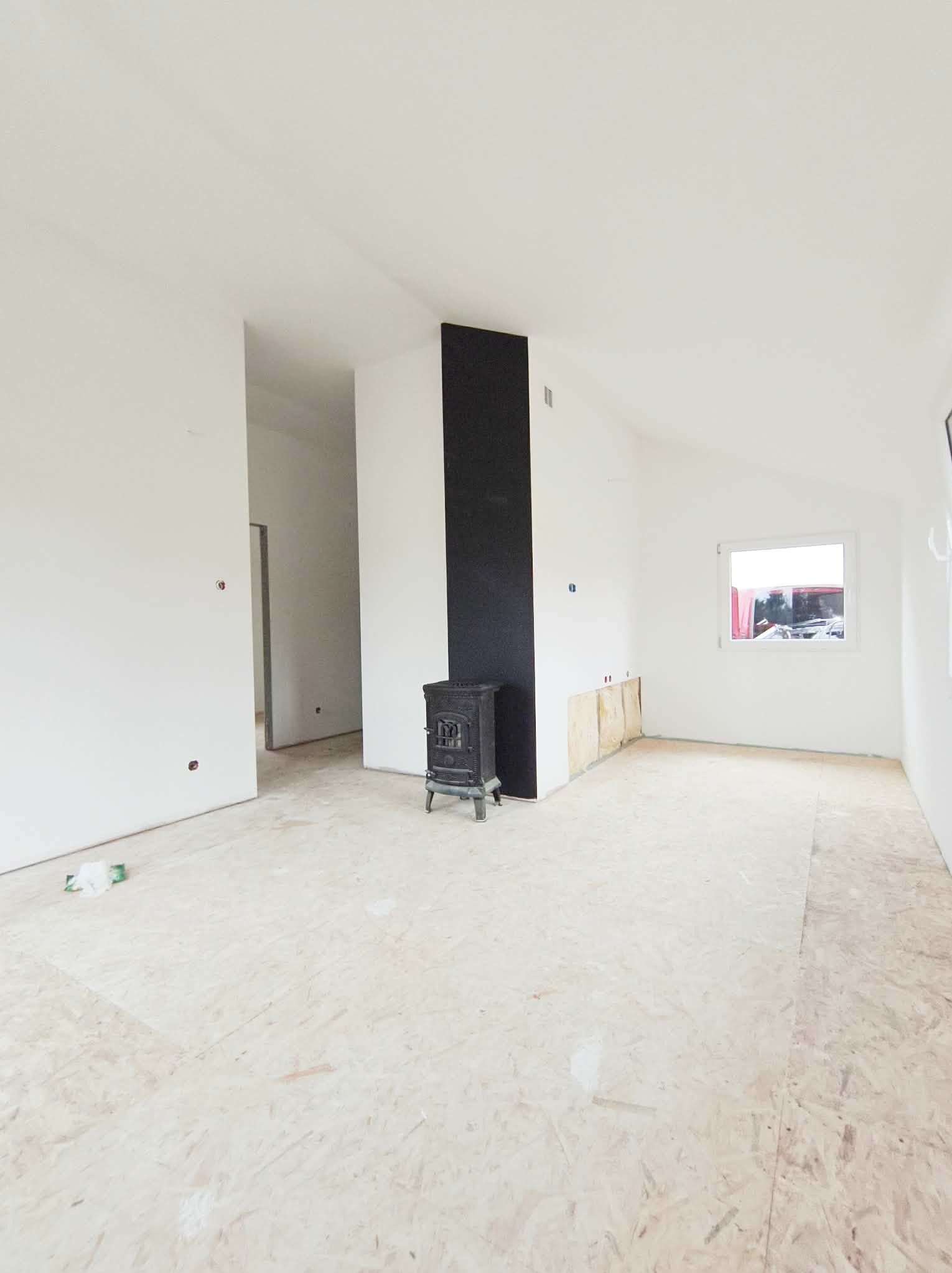Wnętrze w trakcie wykańczania: białe ściany, surowa podłoga OSB, czarny element dekoracyjny kominka i okno. Widoczne ślady po instalacjach.