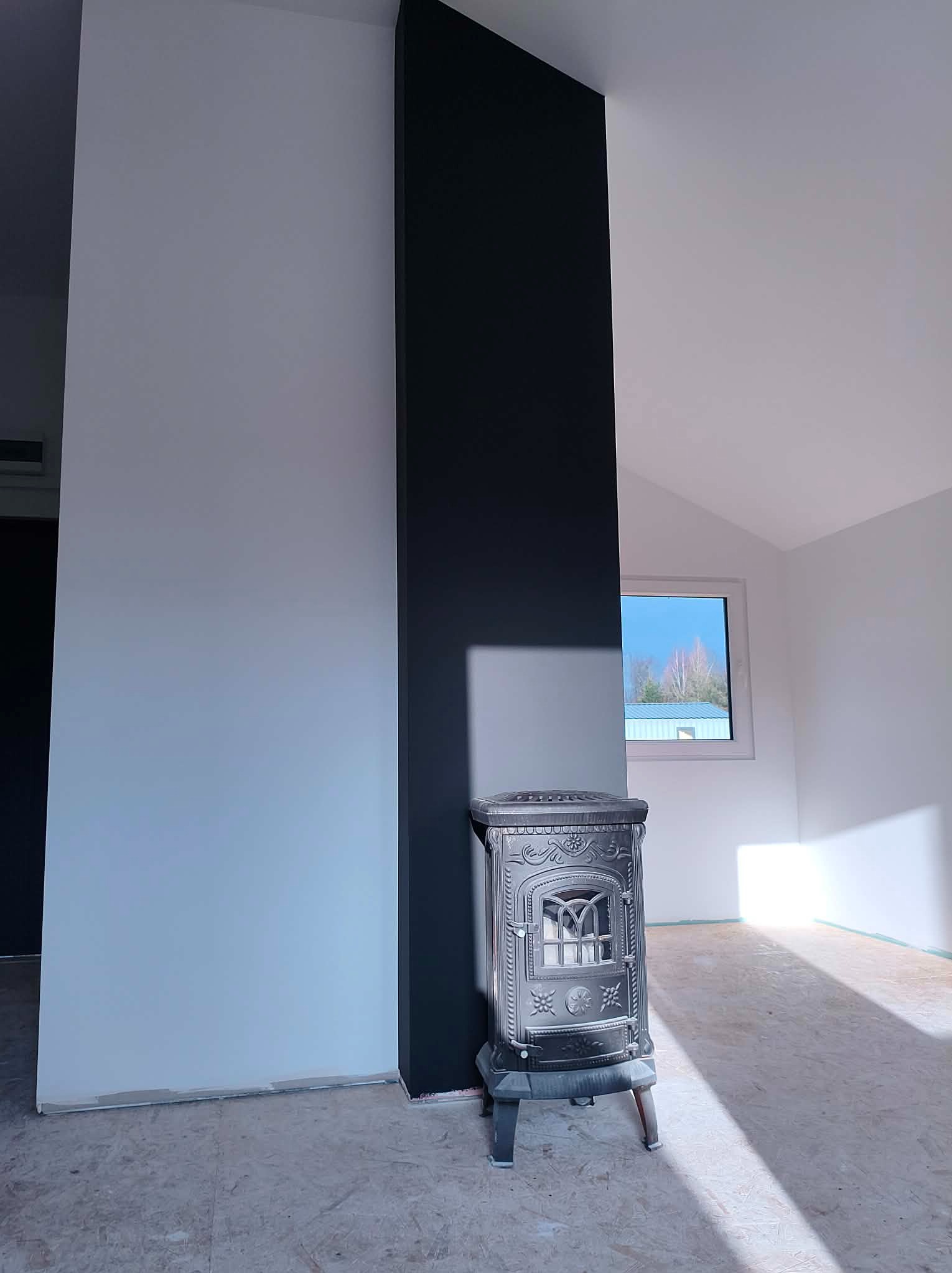 Wnętrze po remoncie: białe ściany, czarny filar, ozdobny piec żeliwny. Podłoga przygotowana do dalszych prac wykończeniowych. Okno z widokiem na zewnątrz.