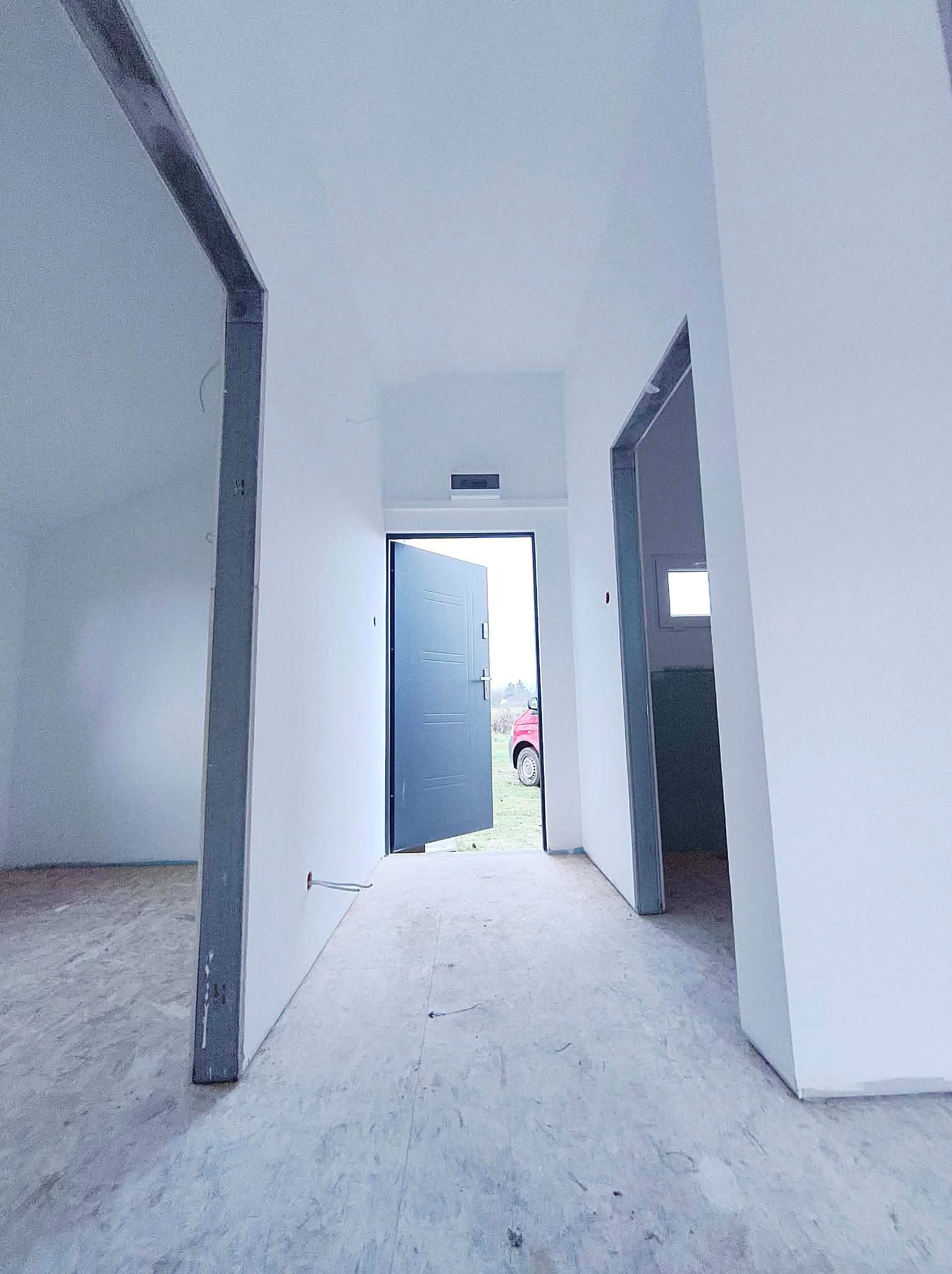 Wnętrze w trakcie wykańczania: białe ściany, surowa podłoga, metalowe ościeżnice, otwarte drzwi wejściowe z widokiem na zewnątrz. Widoczne kable elektryczne.