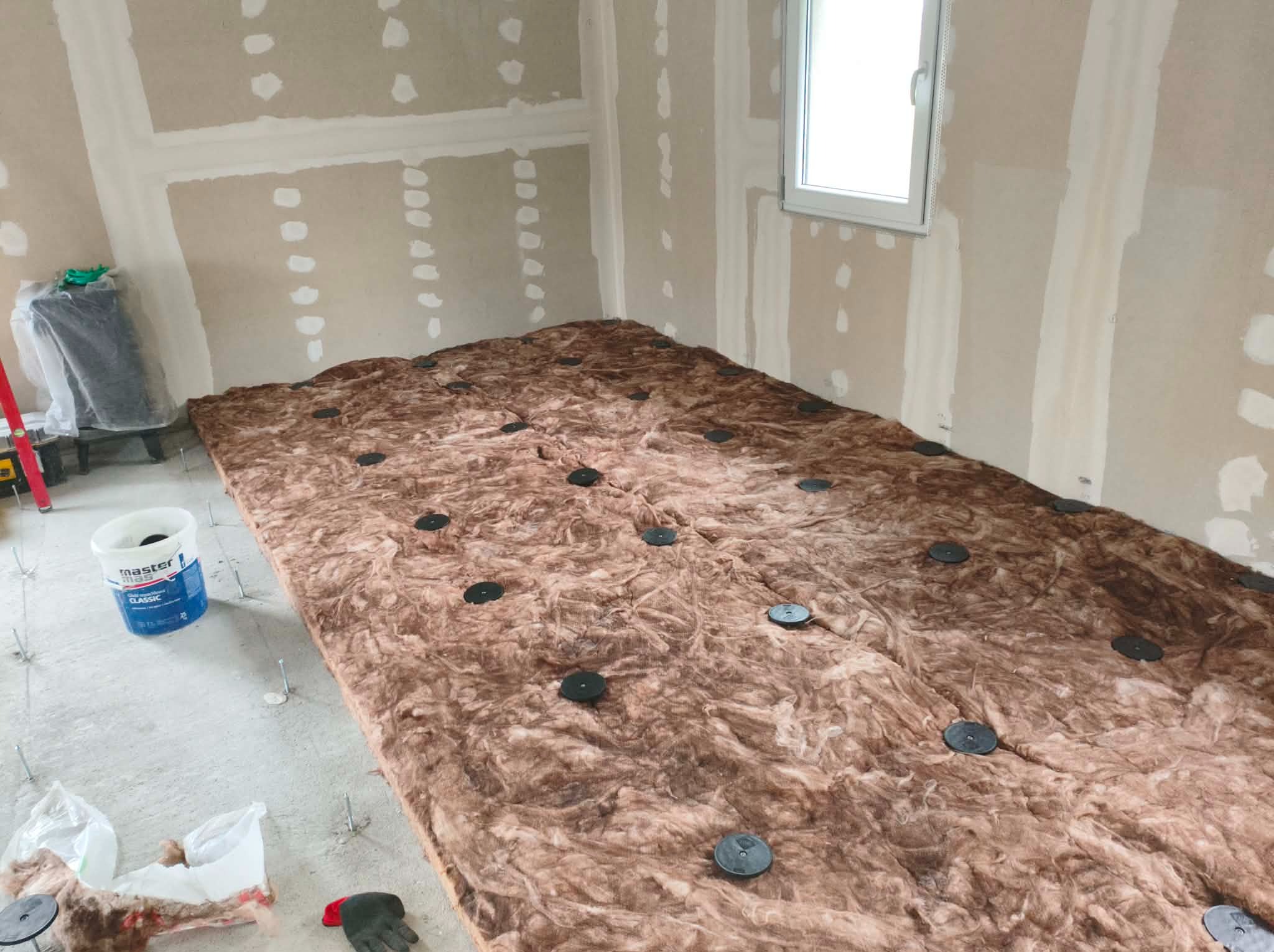 Wnętrze w trakcie prac: wełna mineralna z czarnymi mocowaniami na podłodze, ściany z płyt kartonowo-gipsowych przed wykończeniem, okno. Widoczny pojemnik z klejem.