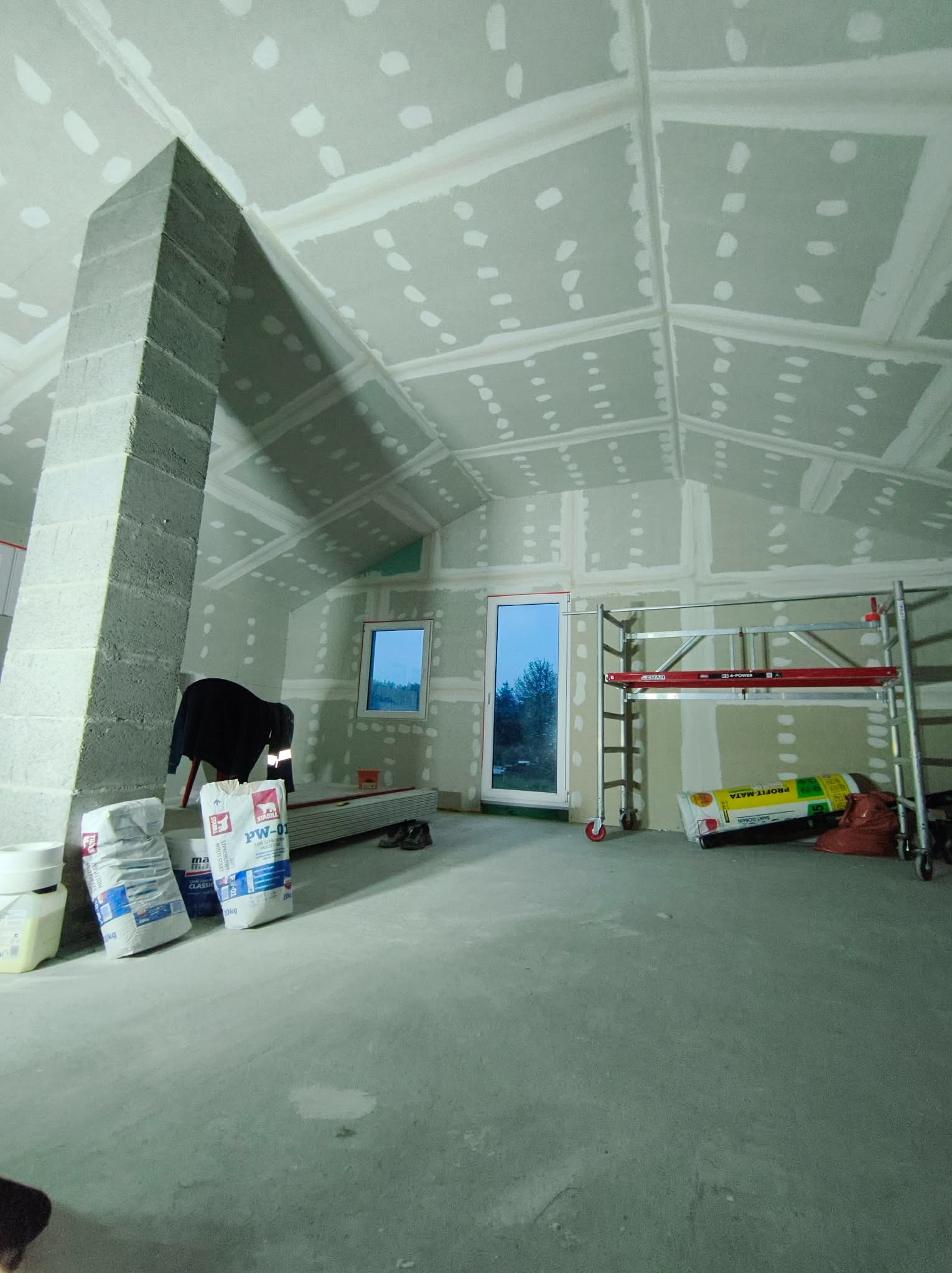 Poddasze w trakcie wykańczania: zamontowane płyty gipsowe na skosach i ścianach, widoczne szpachlowane łączenia, rusztowanie, worki z zaprawą. Prace wykończeniowe.