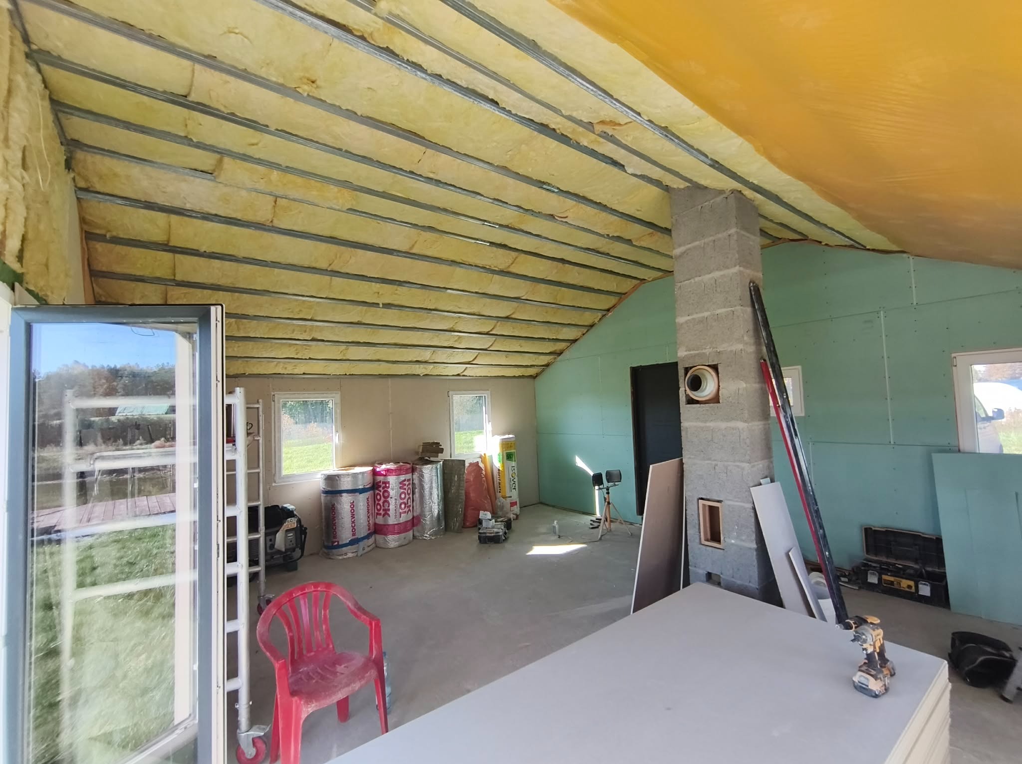 Poddasze w trakcie ocieplania wełną mineralną, widoczne metalowe profile, płyty gipsowe i narzędzia. Przez okno widać zieleń. Charakterystyczny komin z pustaków.