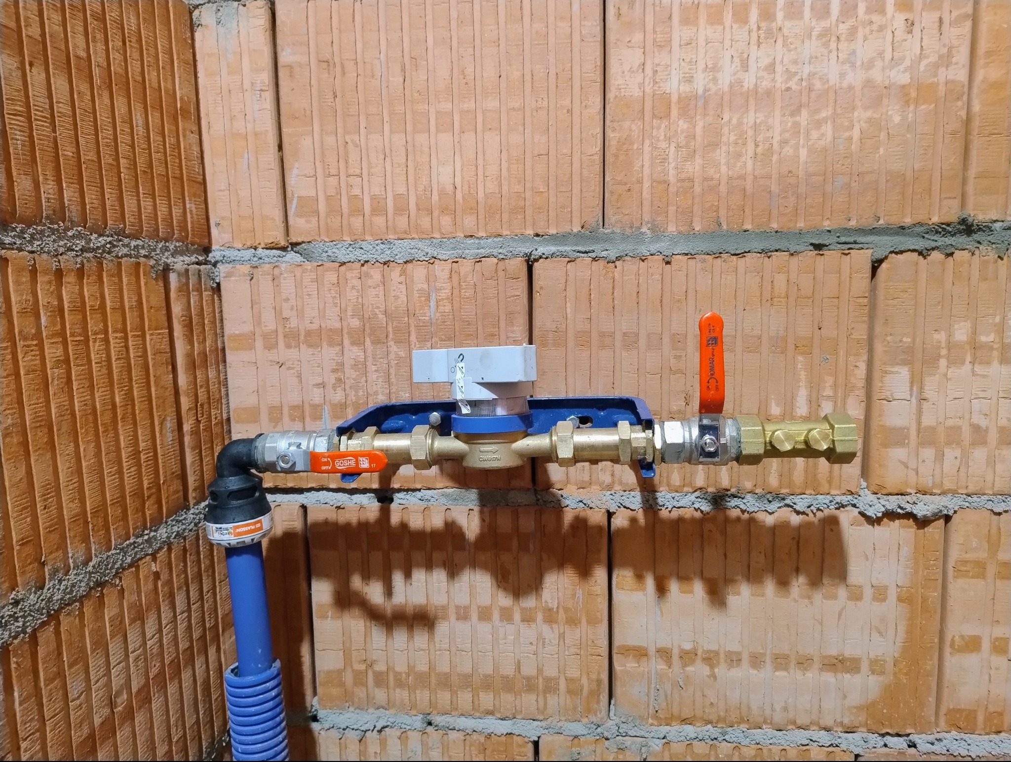 Zamontowany wodomierz z zaworami na ścianie z cegły kratówki. Widoczny fragment niebieskiej rury doprowadzającej wodę. Instalacja wodna w surowym stanie.
