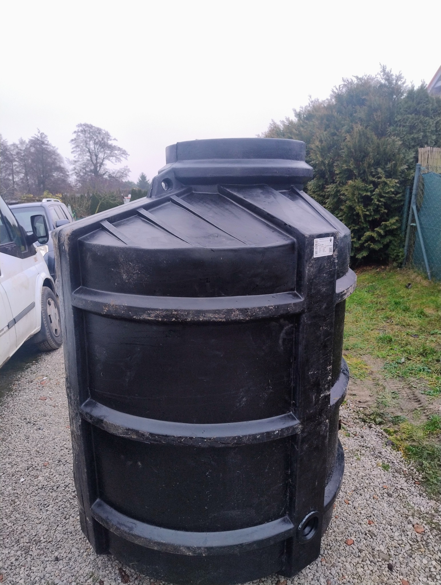 Czarny, plastikowy zbiornik na wodę deszczową z widocznymi przetłoczeniami, stojący na żwirowym podłożu, z fragmentem samochodu i zieleni w tle.