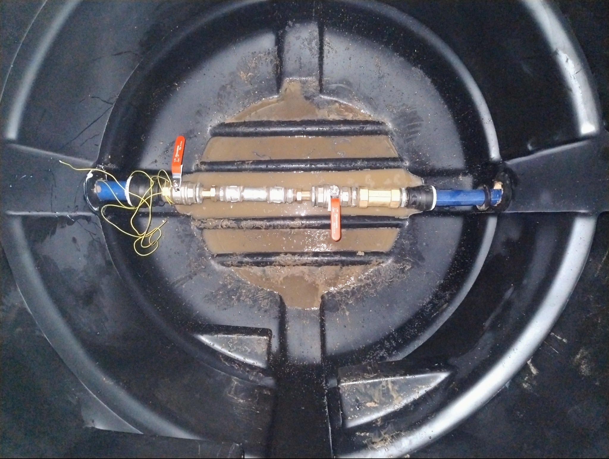 Wnętrze czarnego zbiornika z instalacją hydrauliczną, widoczne rury, zawory z pomarańczowymi uchwytami i żółty przewód. Część zbiornika wypełniona wodą.