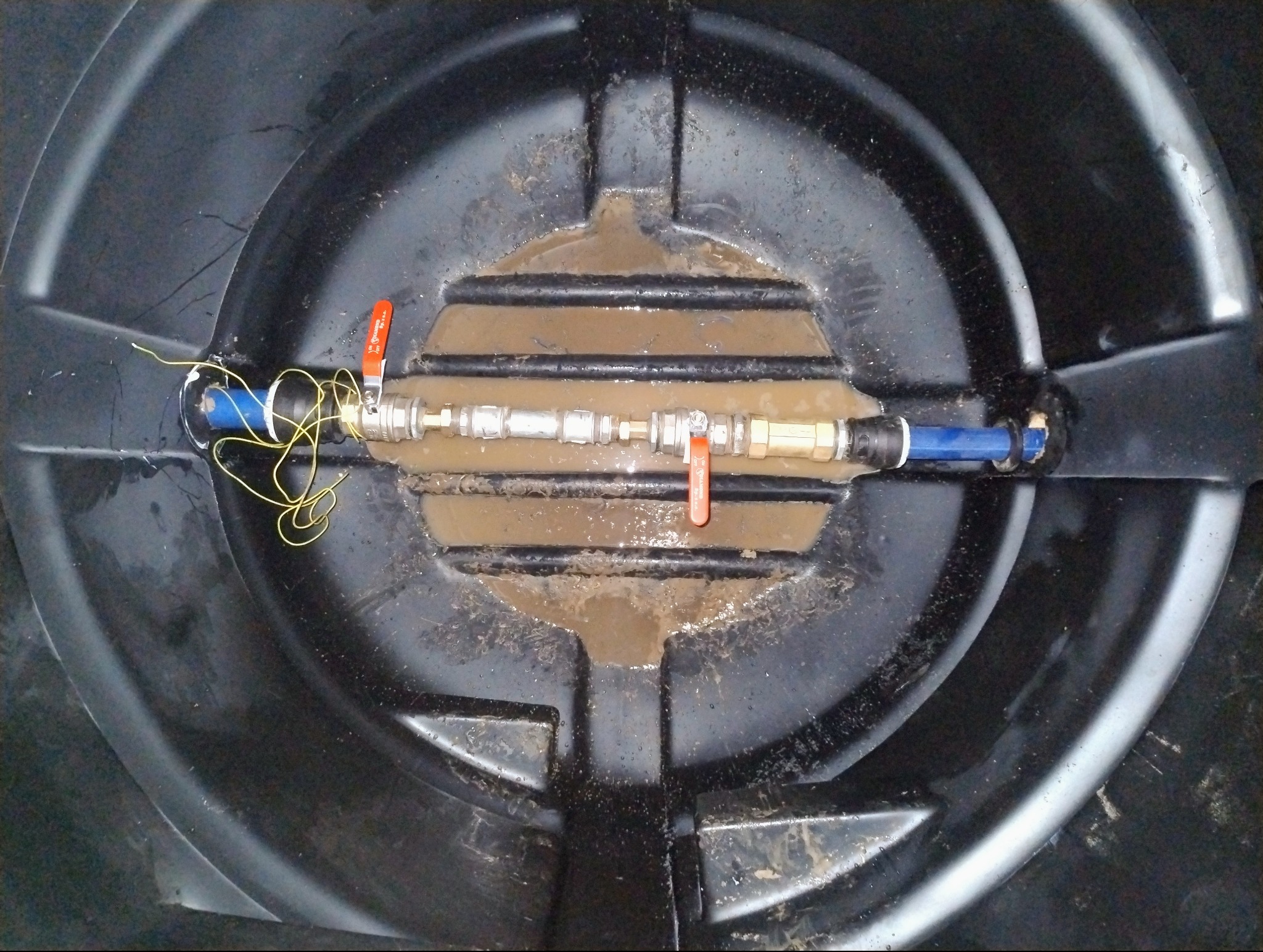 Wnętrze czarnego zbiornika z podłączeniem hydraulicznym, widok z góry. Widoczne rury, zawory z czerwoną rączką i żółte przewody. Woda w zbiorniku.