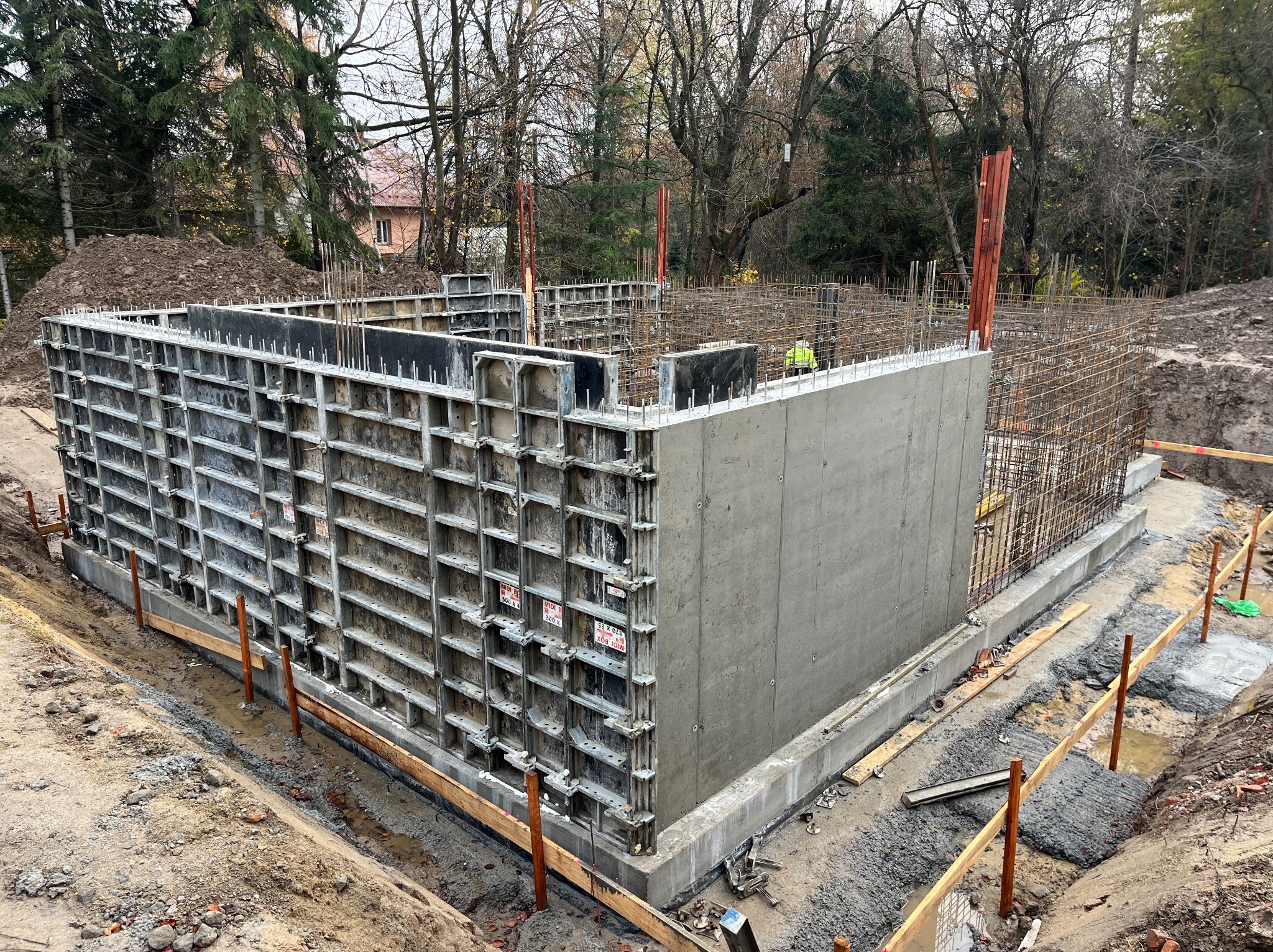 Wylany fundament z szalunkiem systemowym i zbrojeniem, widok z góry. Etap budowy domu, teren budowy z drzewami w tle. Prace fundamentowe w toku.