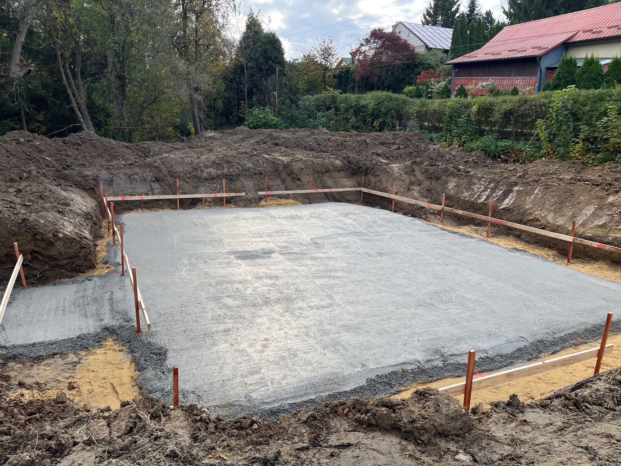 Wylany fundament pod budowę domu, widoczne drewniane szalunki i przygotowany teren. W tle zieleń drzew i zabudowania mieszkalne.