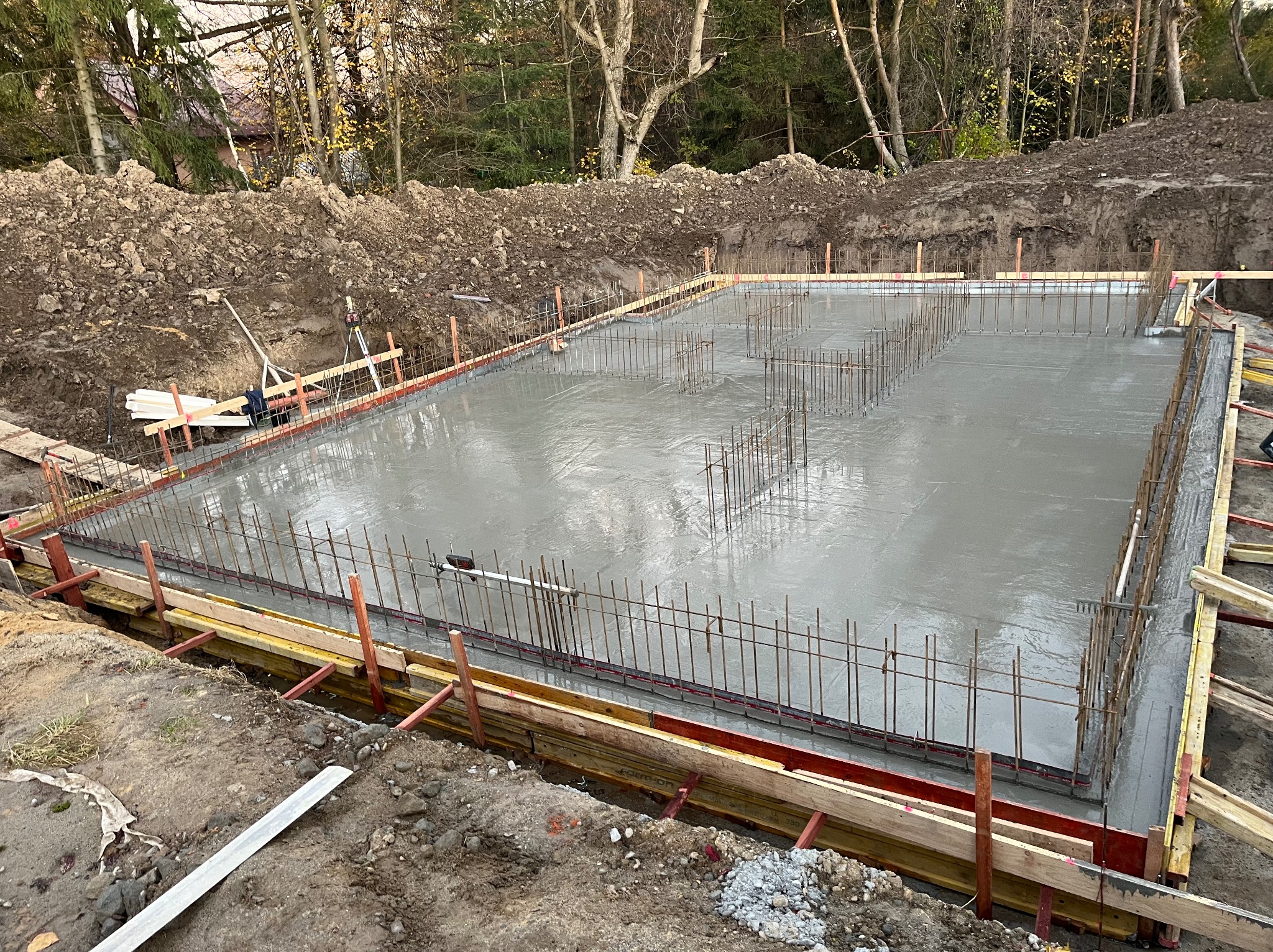 Zalany betonem fundament z wystającymi prętami zbrojeniowymi, otoczony drewnianym szalunkiem. W tle wykopana ziemia i drzewa.