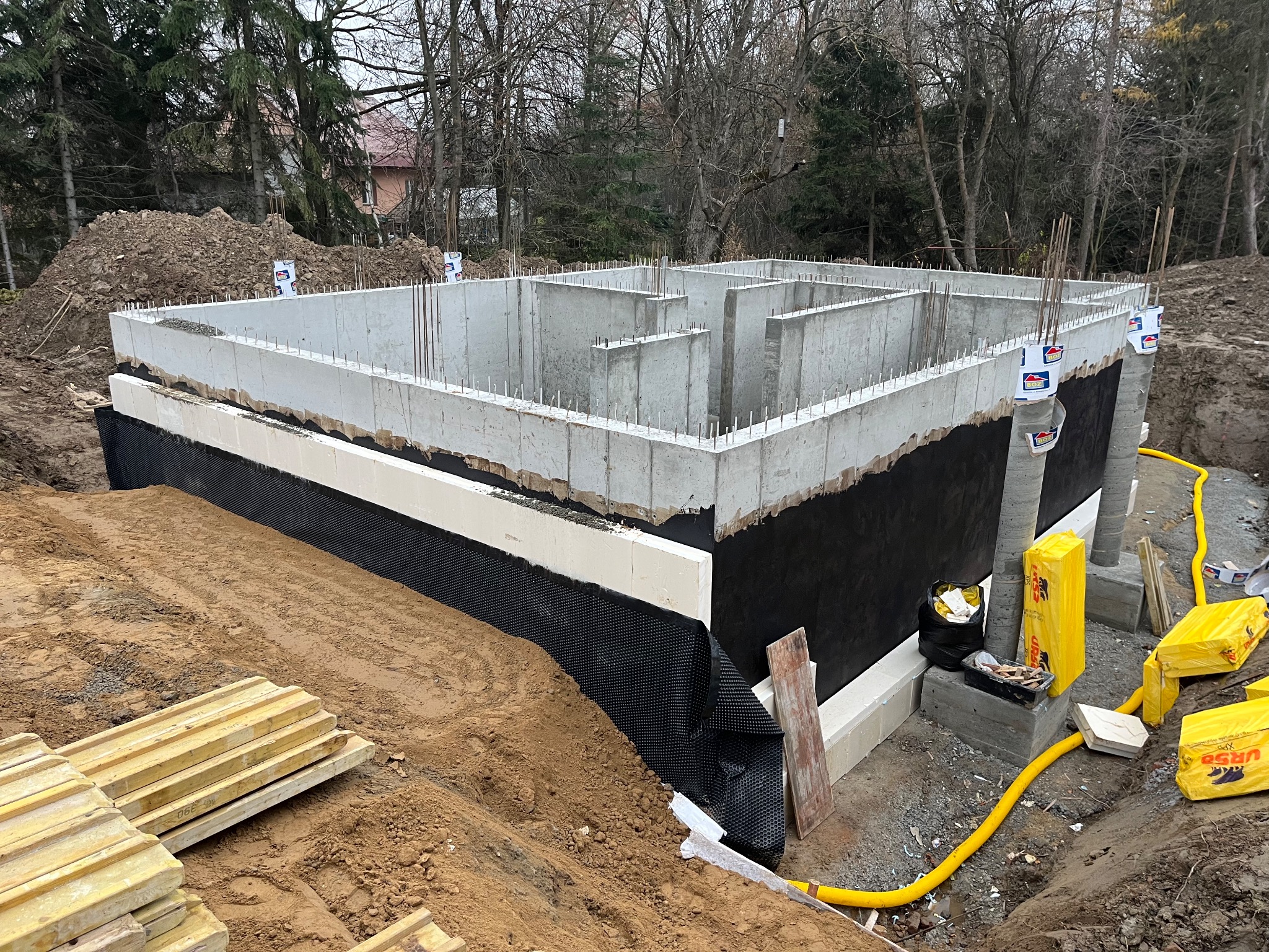 Zabezpieczony fundament z izolacją pionową, styrodurem i folią kubełkową. Widoczne zbrojenia i betonowe ściany. Żółte rury wentylacyjne i paczki izolacji termicznej.