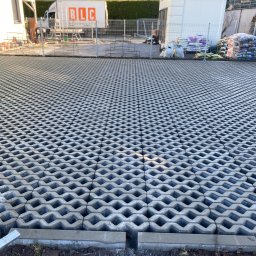 Uslugi kamieniarsko -brukarskie - Ułożona szara ażurowa kostka brukowa na podjeździe, widok z góry. W tle budynek, samochód dostawczy i worki z materiałami budowlanymi.