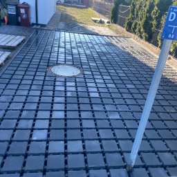 Uslugi kamieniarsko -brukarskie - Ułożona, grafitowa kostka brukowa z kratką drenażową i studzienką kanalizacyjną. W tle zieleń i zabudowa. Widoczny słupek z oznaczeniem wodociągu.