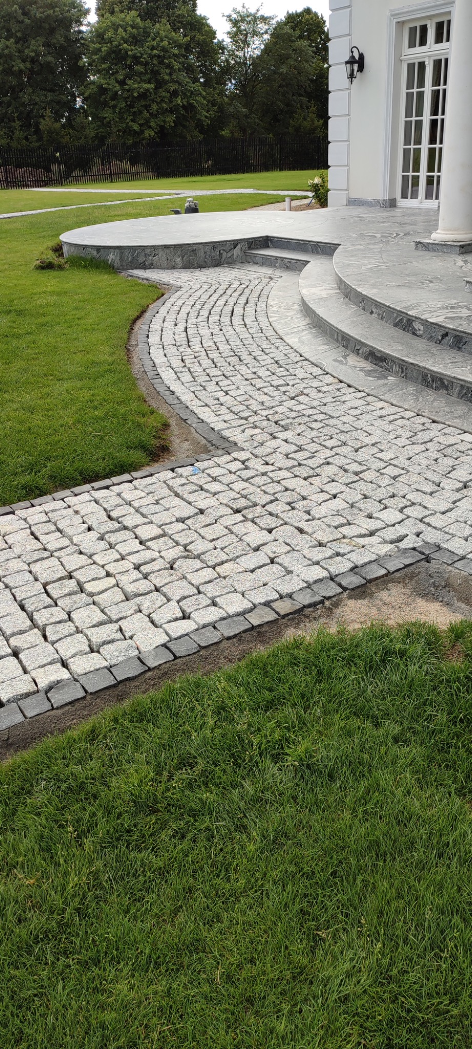 Zakrzywiona ścieżka z jasnoszarej kostki granitowej, obrzeża z ciemniejszej kostki, prowadząca do schodów i wejścia do domu z kolumnami. Zadbane trawniki po bokach.