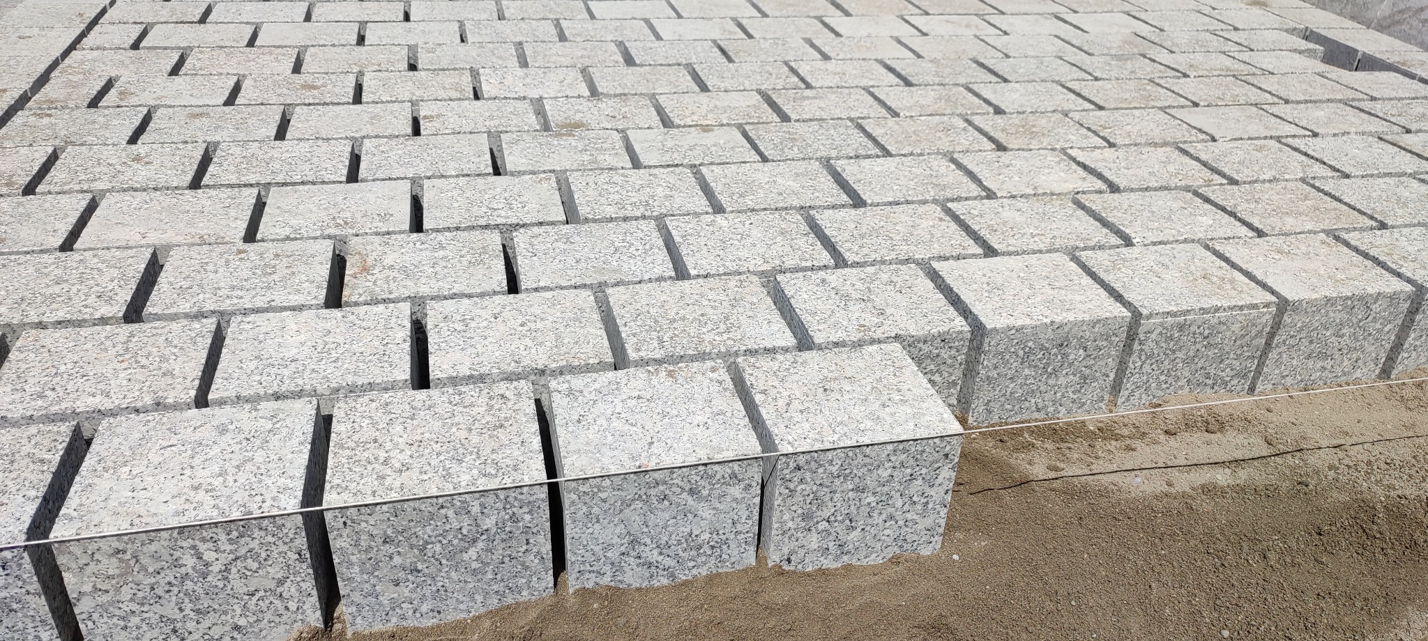 Jasnoszara kostka granitowa ułożona w rzędach, widoczne spoiny i linia wyznaczająca poziom. Surowy piasek w tle. Delikatne światło podkreśla fakturę kamienia.
