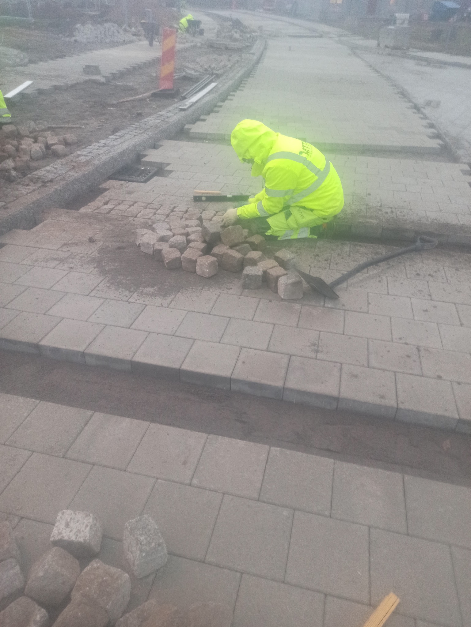Pracownik w odblaskowym stroju układa kostkę granitową na chodniku, używając poziomicy. W tle widoczne inne osoby i teren budowy.