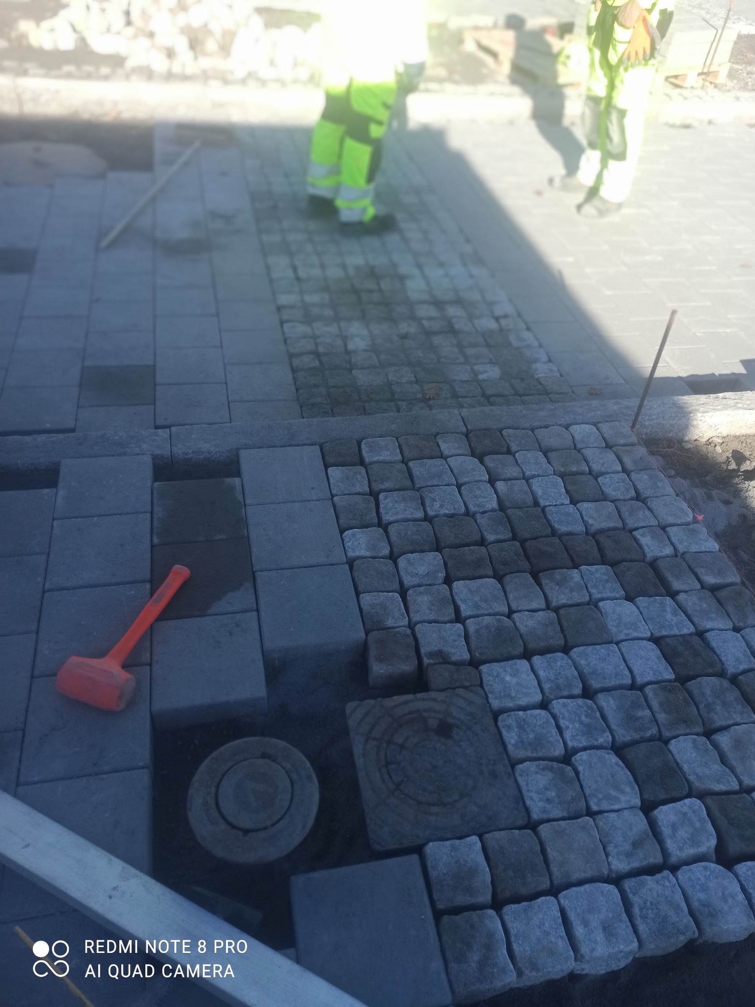 Fragment układanej nawierzchni z kostki granitowej w odcieniach szarości, z widocznym fragmentem studzienki i gumowym młotkiem. W tle pracownicy w odblaskowych ubraniach.