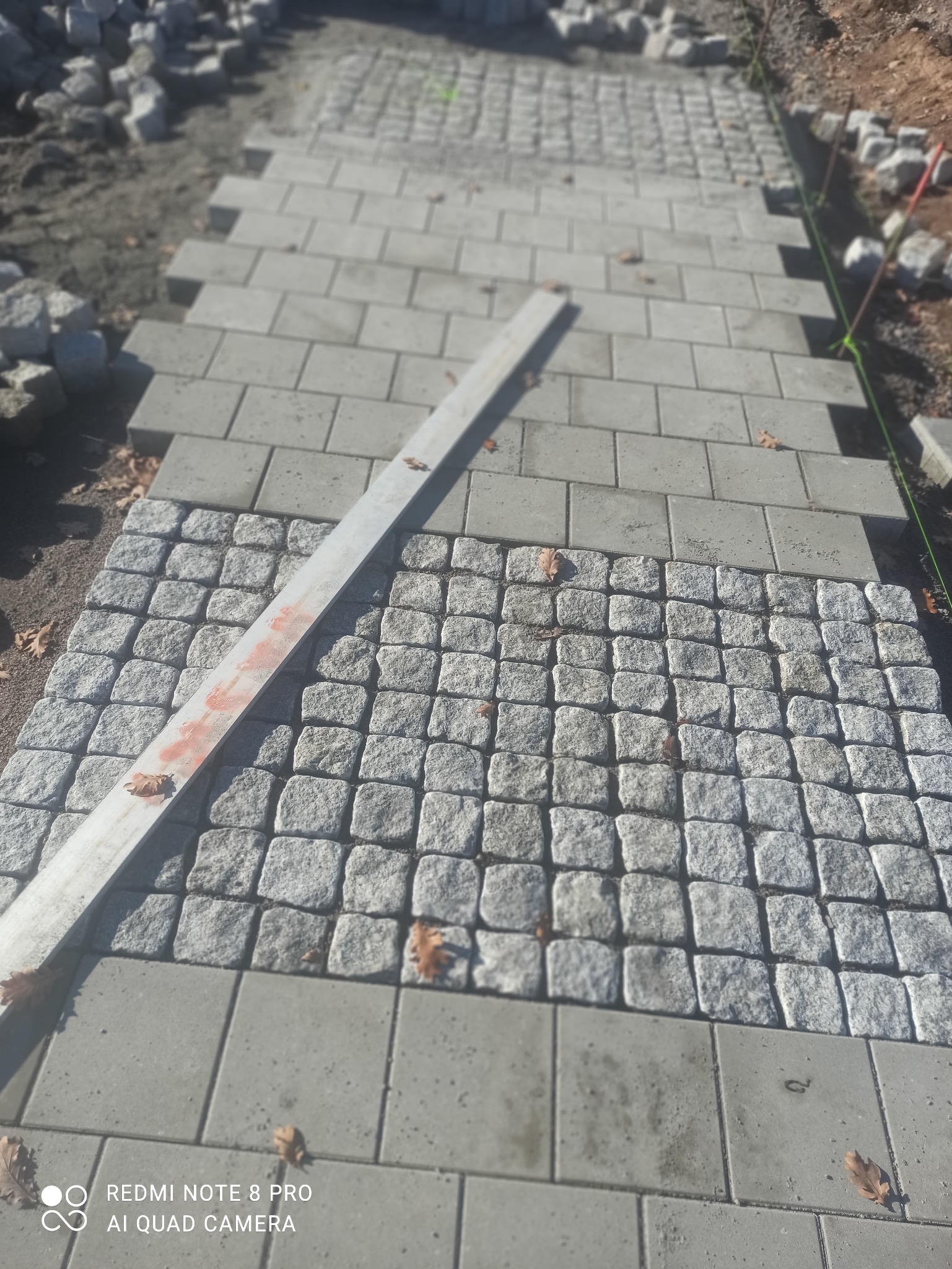 Fragment ścieżki z kostki granitowej i brukowej w trakcie realizacji. Widoczna poziomica i zapas kostki. Jesienne liście na powierzchni.